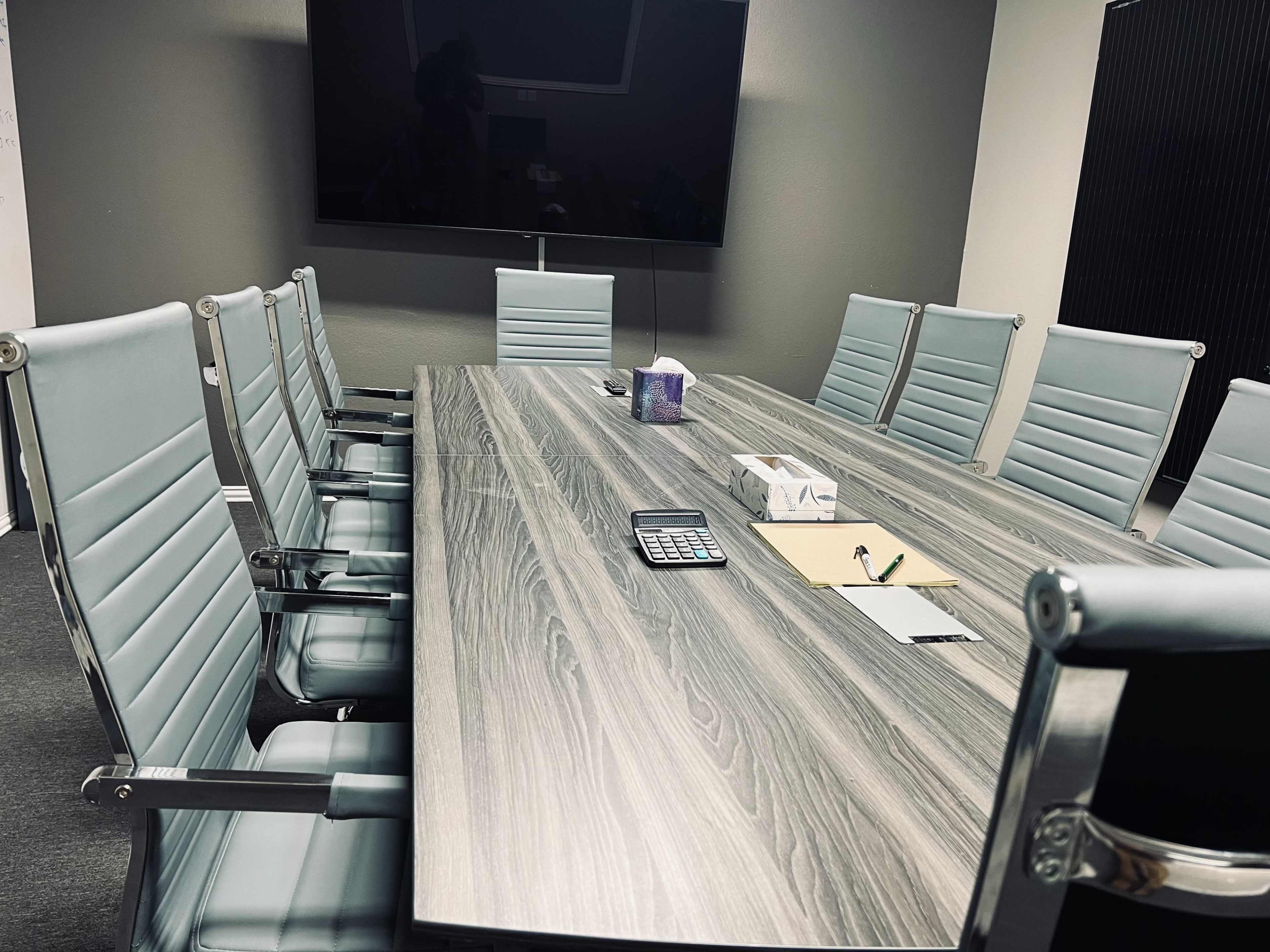 A conference room with a long wooden table, eight light blue chairs, and a large screen on the wall.