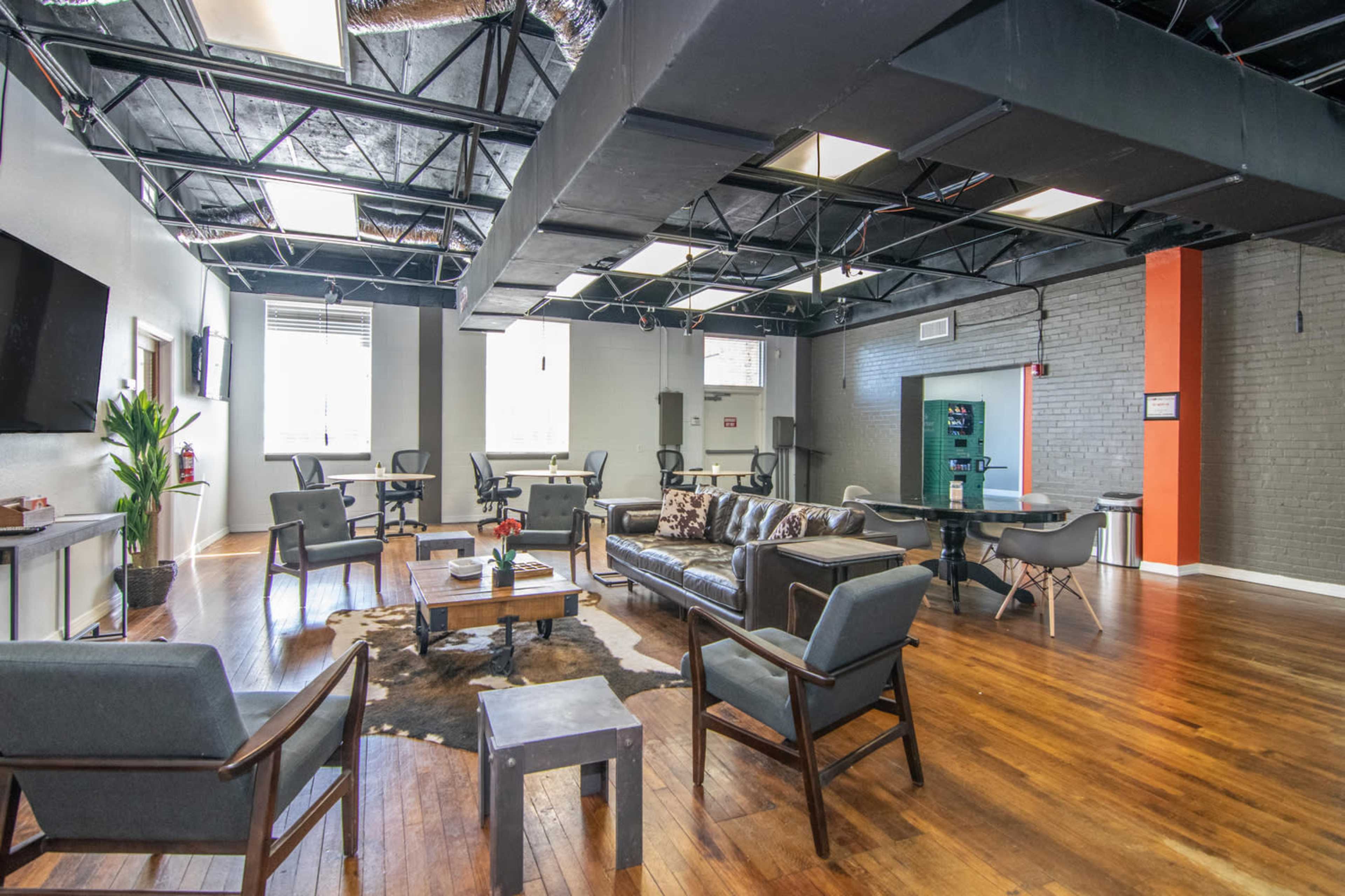 A shared office space with a mix of seating areas, including a couch and several tables, against a backdrop of exposed beams and large windows.