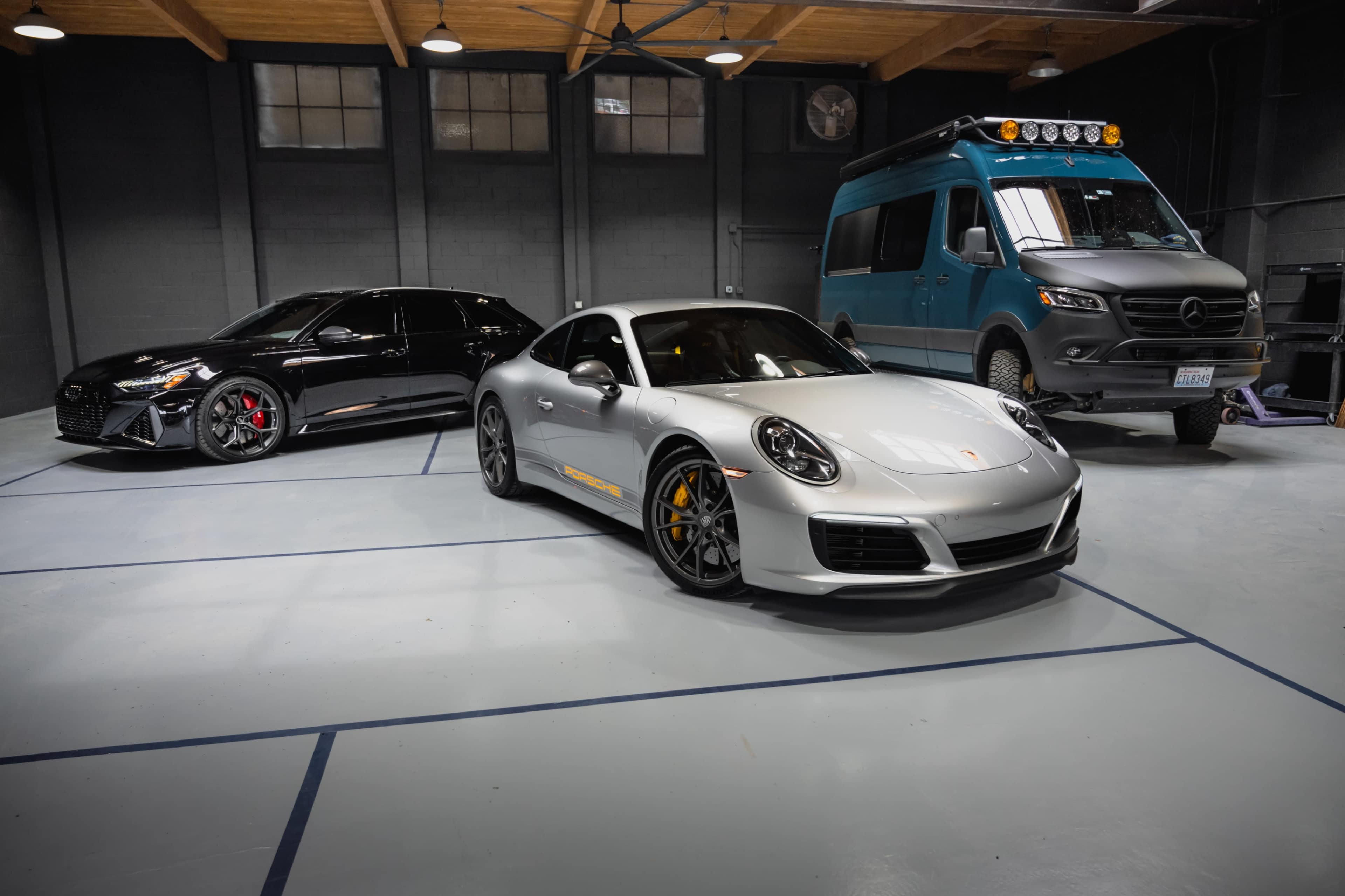 The image shows three vehicles parked in a well-lit garage: a black sedan, a silver sports car, and a blue van with roof lights.