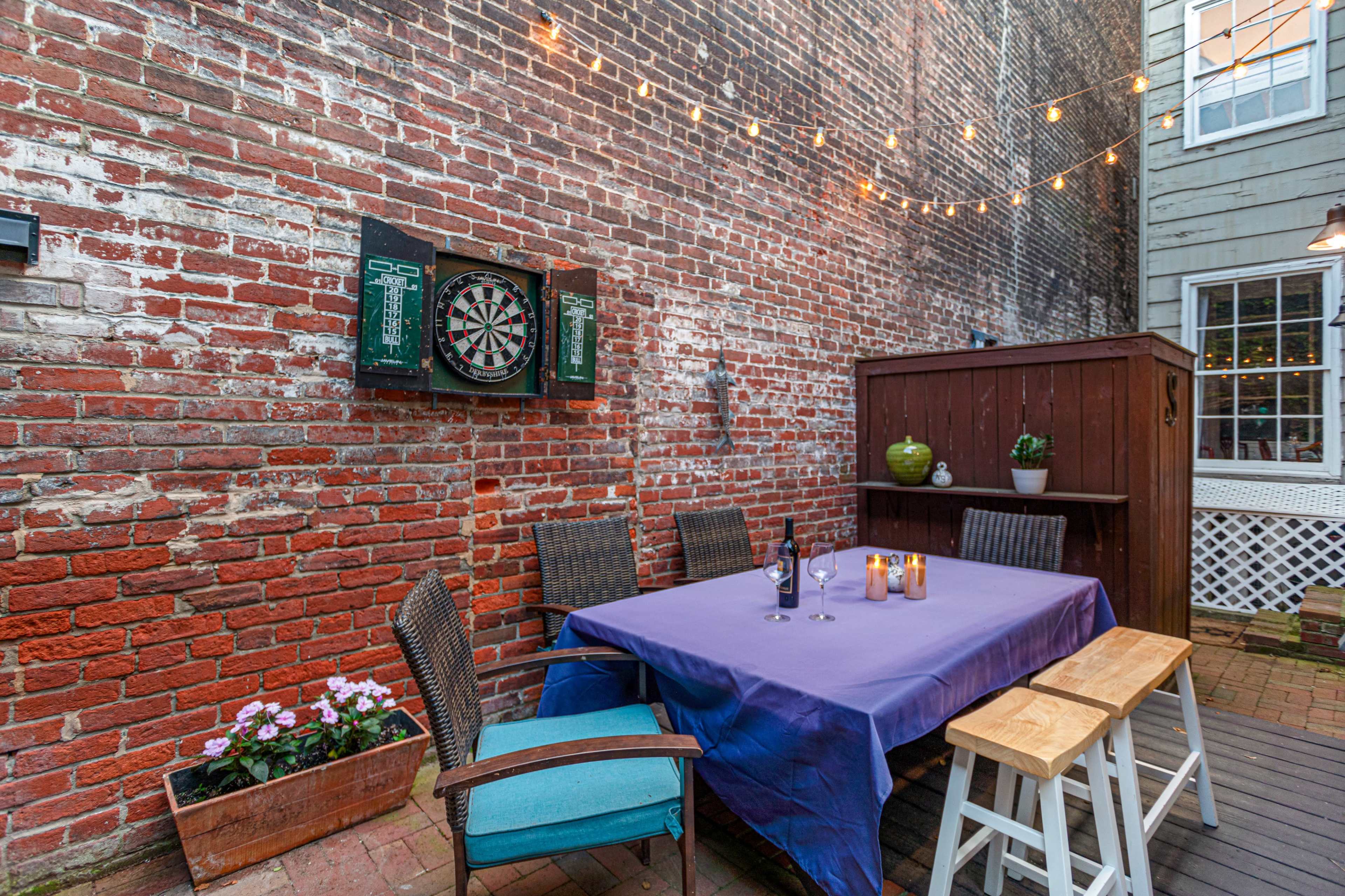 The image shows an outdoor patio area with a brick wall, a large table set with candles, and a dartboard mounted on the wall, illuminated by string lights overhead.