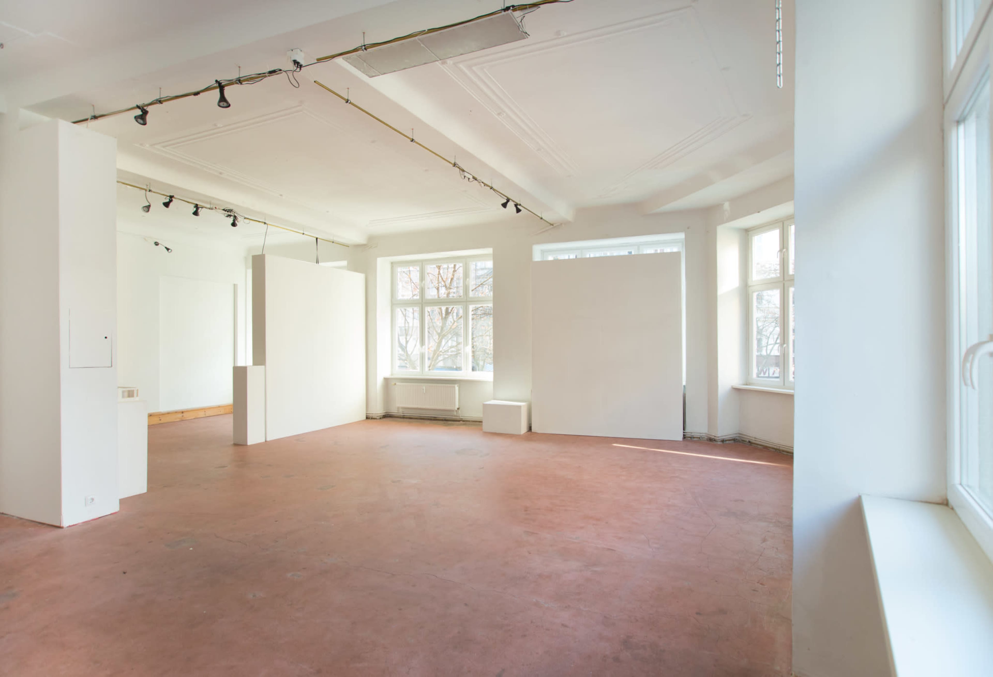 A spacious, empty gallery with white walls, large windows, and a reddish-brown floor.