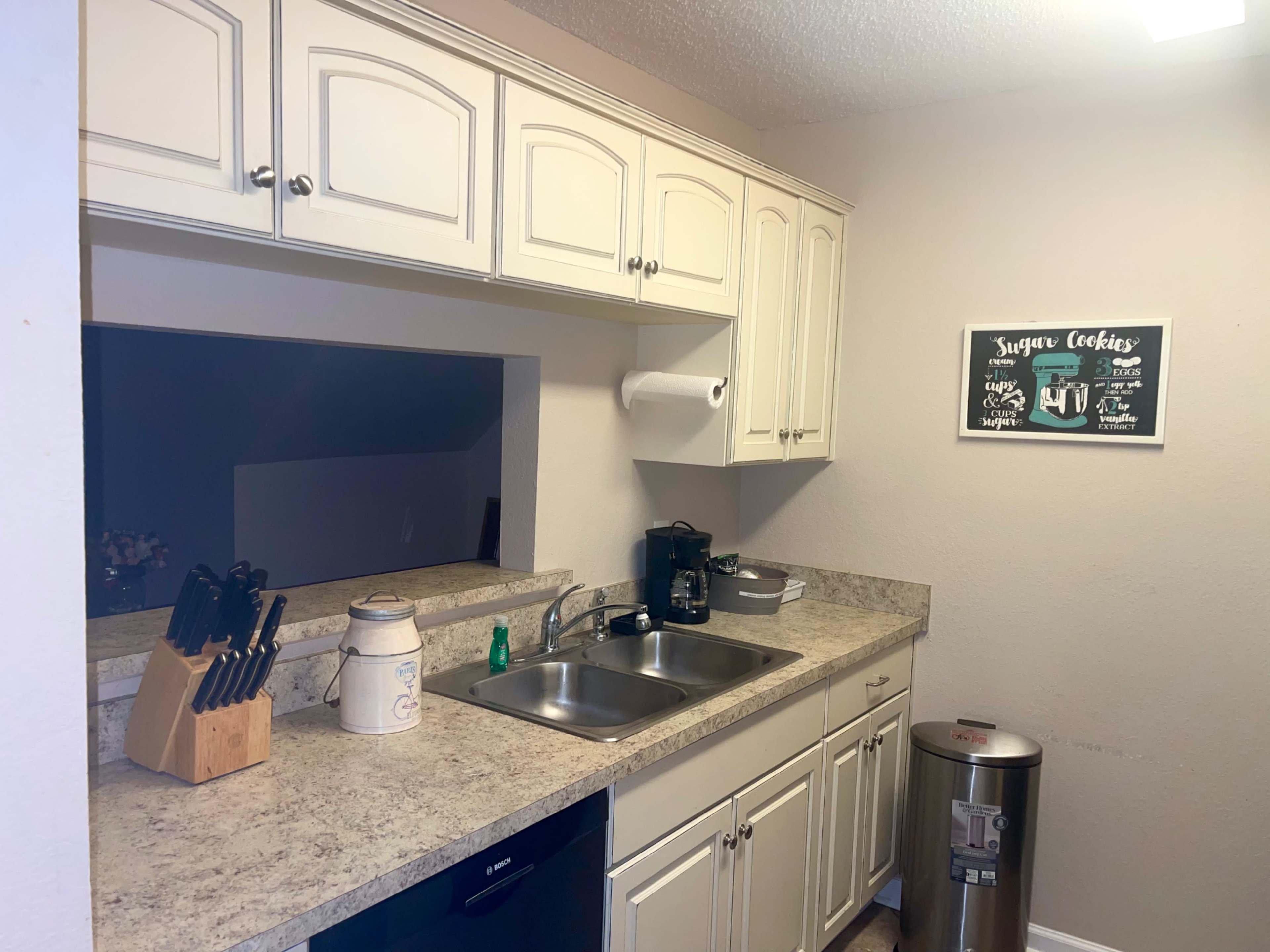 A small kitchen features white cabinetry, a double sink under a window, and various appliances, including a coffee maker and a trash bin.
