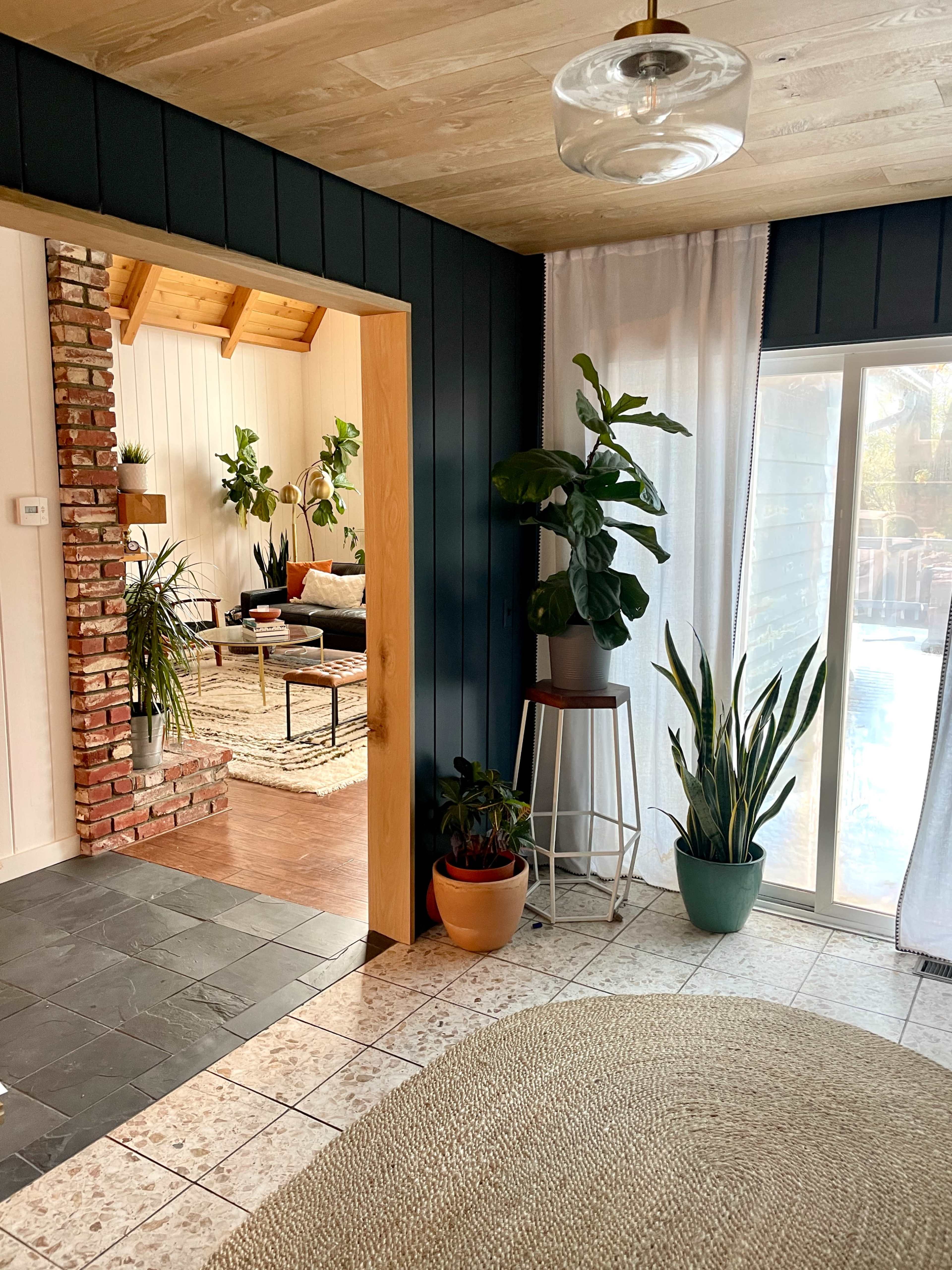 The image shows a cozy interior space featuring a mix of plants, a brick wall, and large windows letting in natural light.