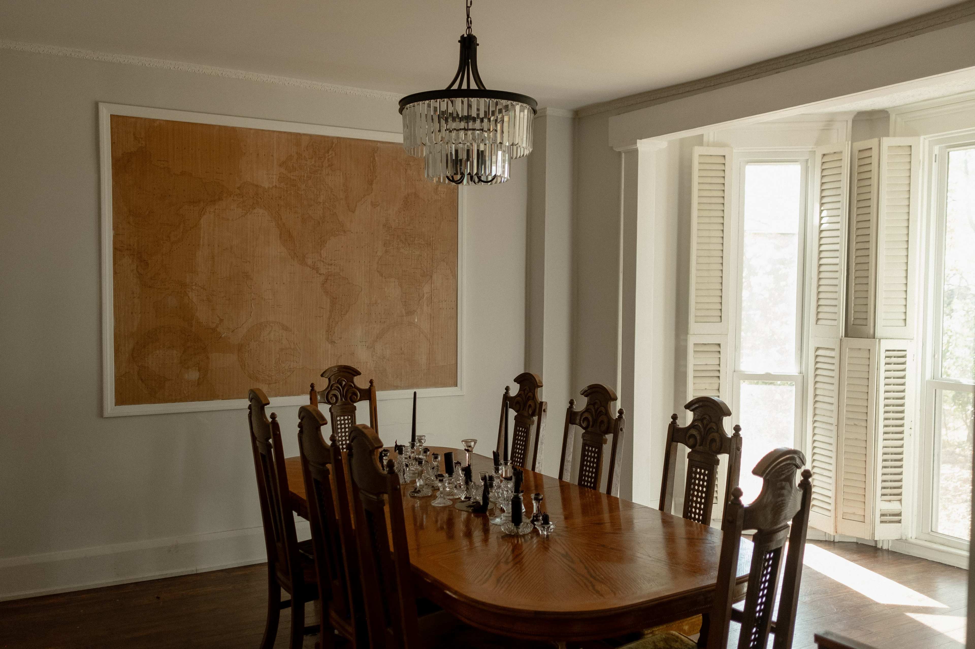 A large wooden dining table with ten chairs is positioned in a room featuring a world map on the wall and windows with shuttered panels.