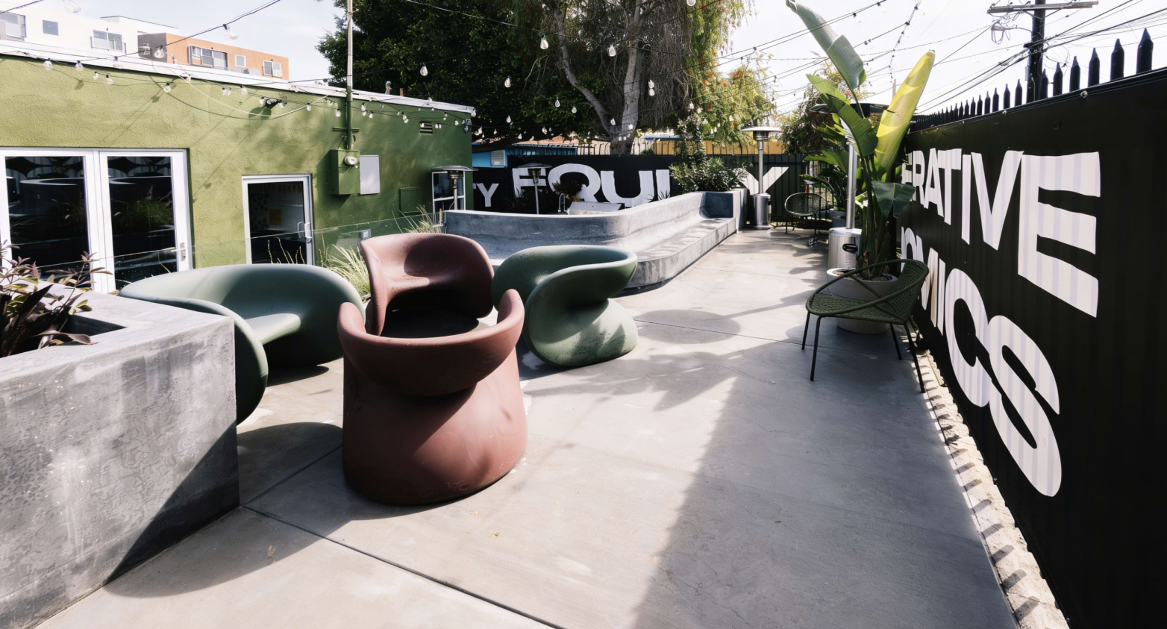 The image shows an outdoor patio area featuring modern seating and a concrete surface, surrounded by greenery and a decorative wall with bold text.