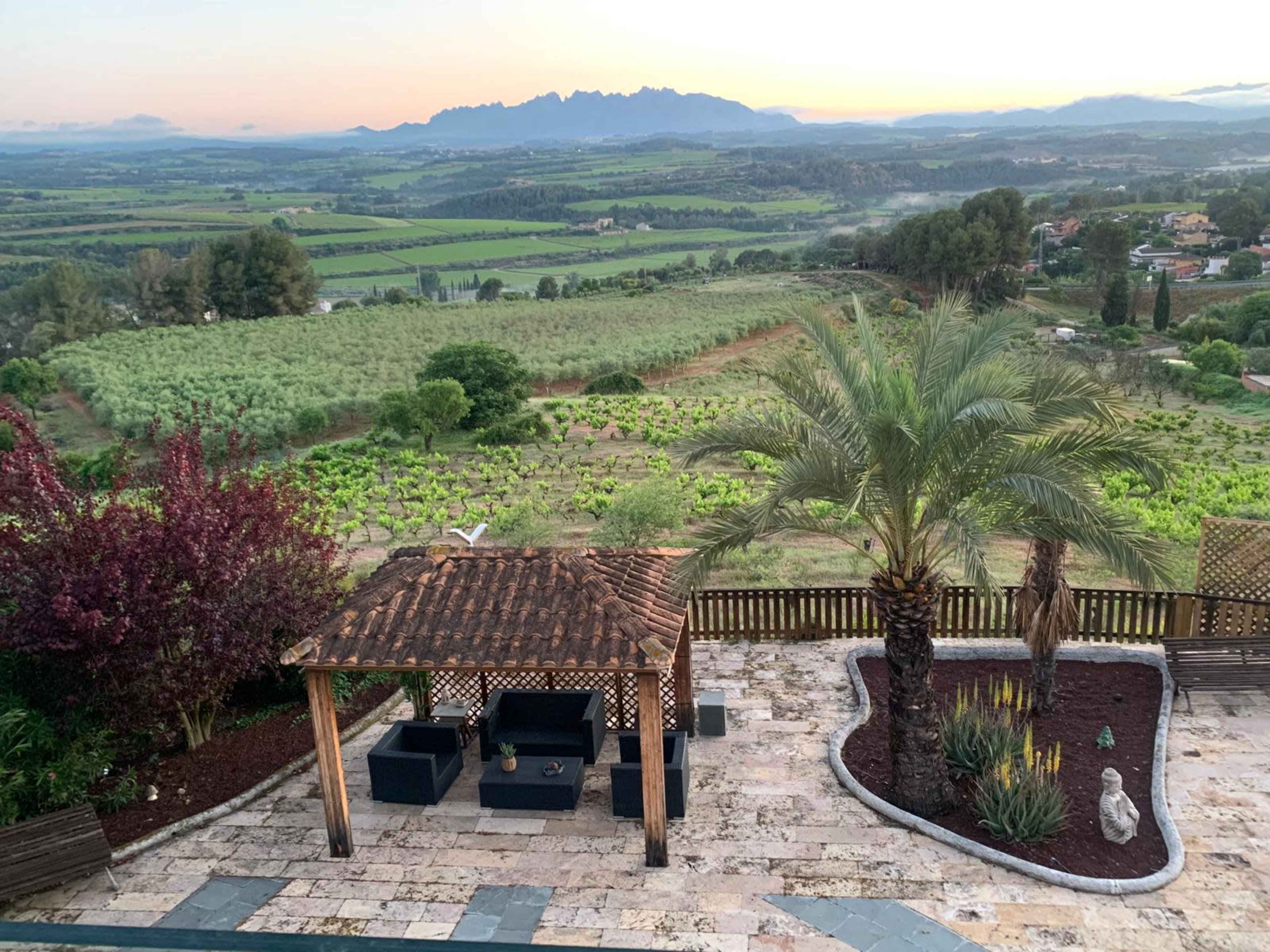 The image shows a landscaped patio with a wooden pergola and seating, overlooking lush green fields and mountains in the background during early morning light.