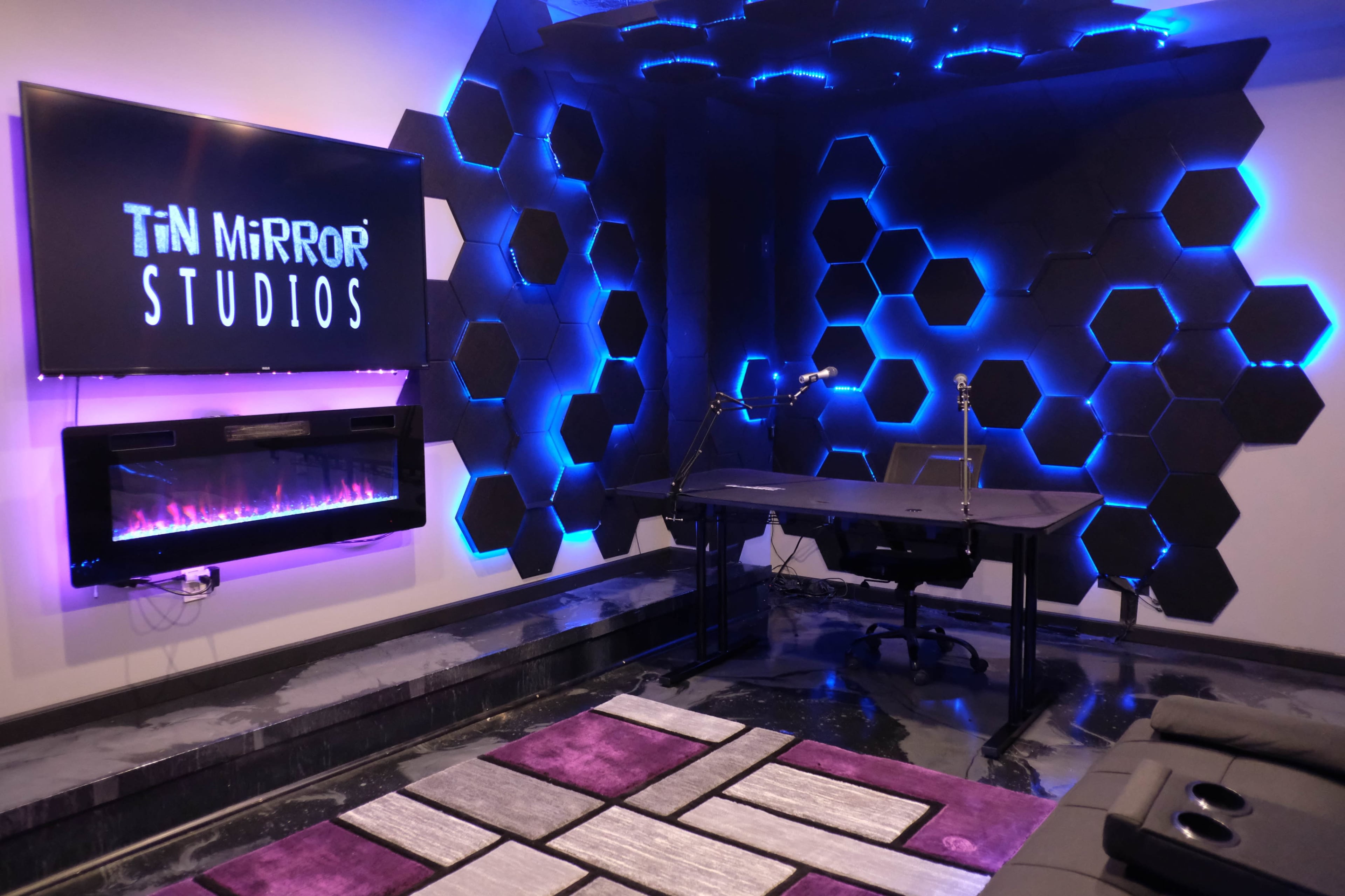 The image shows a modern studio space with a black desk, two microphones, a large screen displaying "TiN MiRROR STUDIOS," and hexagon-shaped wall decor illuminated by blue lighting.