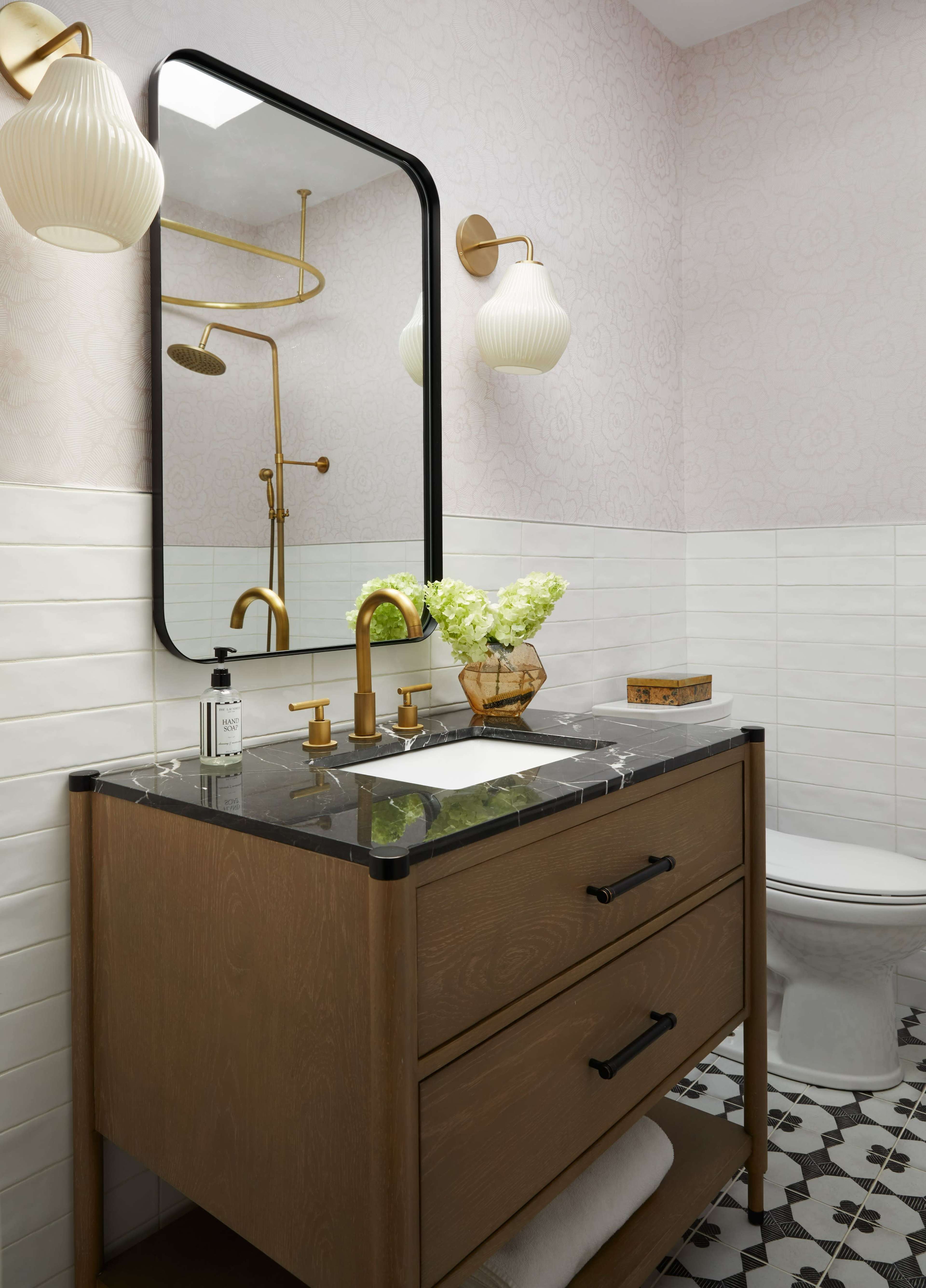 A modern bathroom featuring a wooden vanity with a black countertop, a round mirror, gold fixtures, and patterned floor tiles.