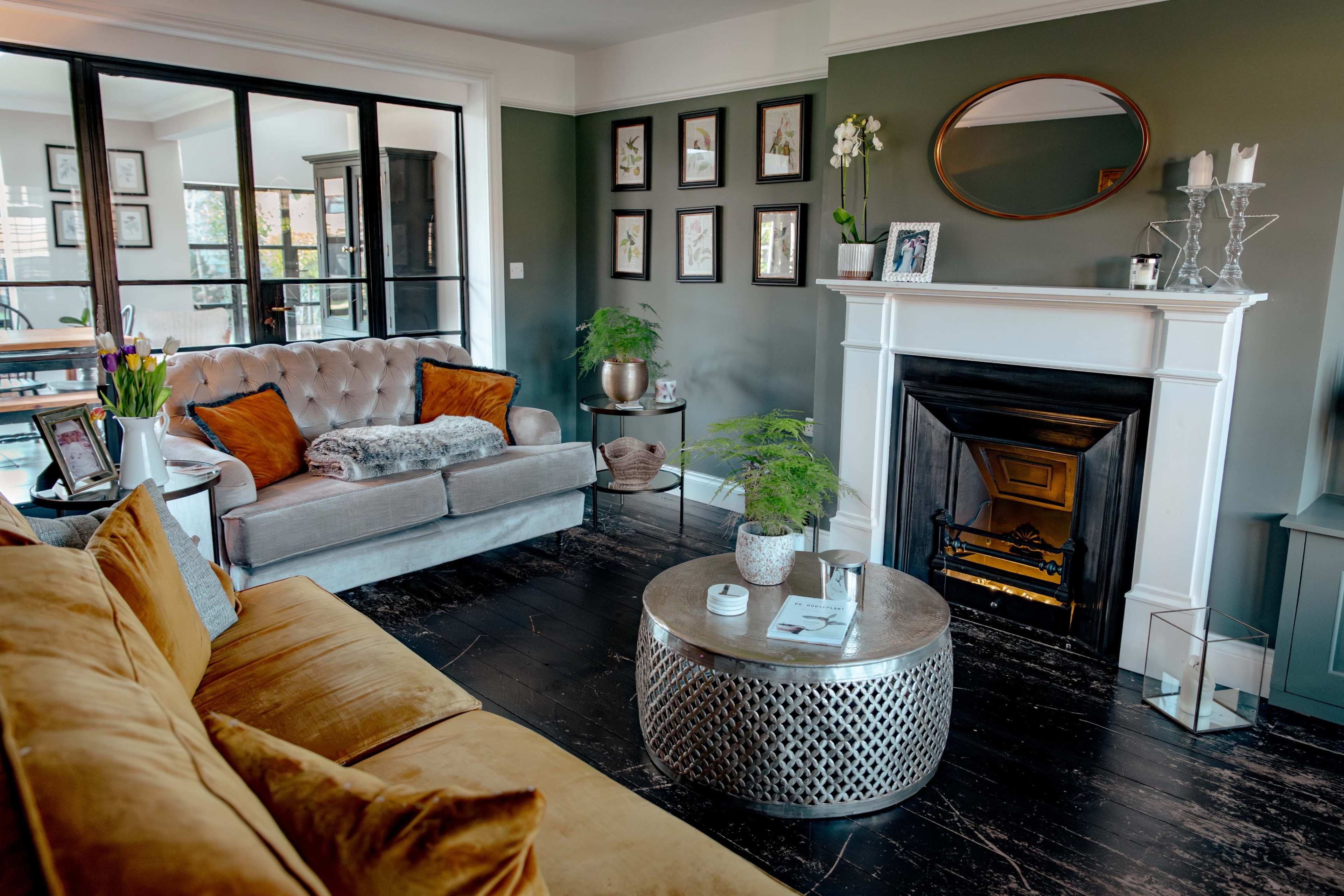 The image shows a well-furnished living room featuring a gray tufted sofa, a mustard-colored sofa, a round coffee table, and a fireplace, all set against a green wall adorned with framed artwork.