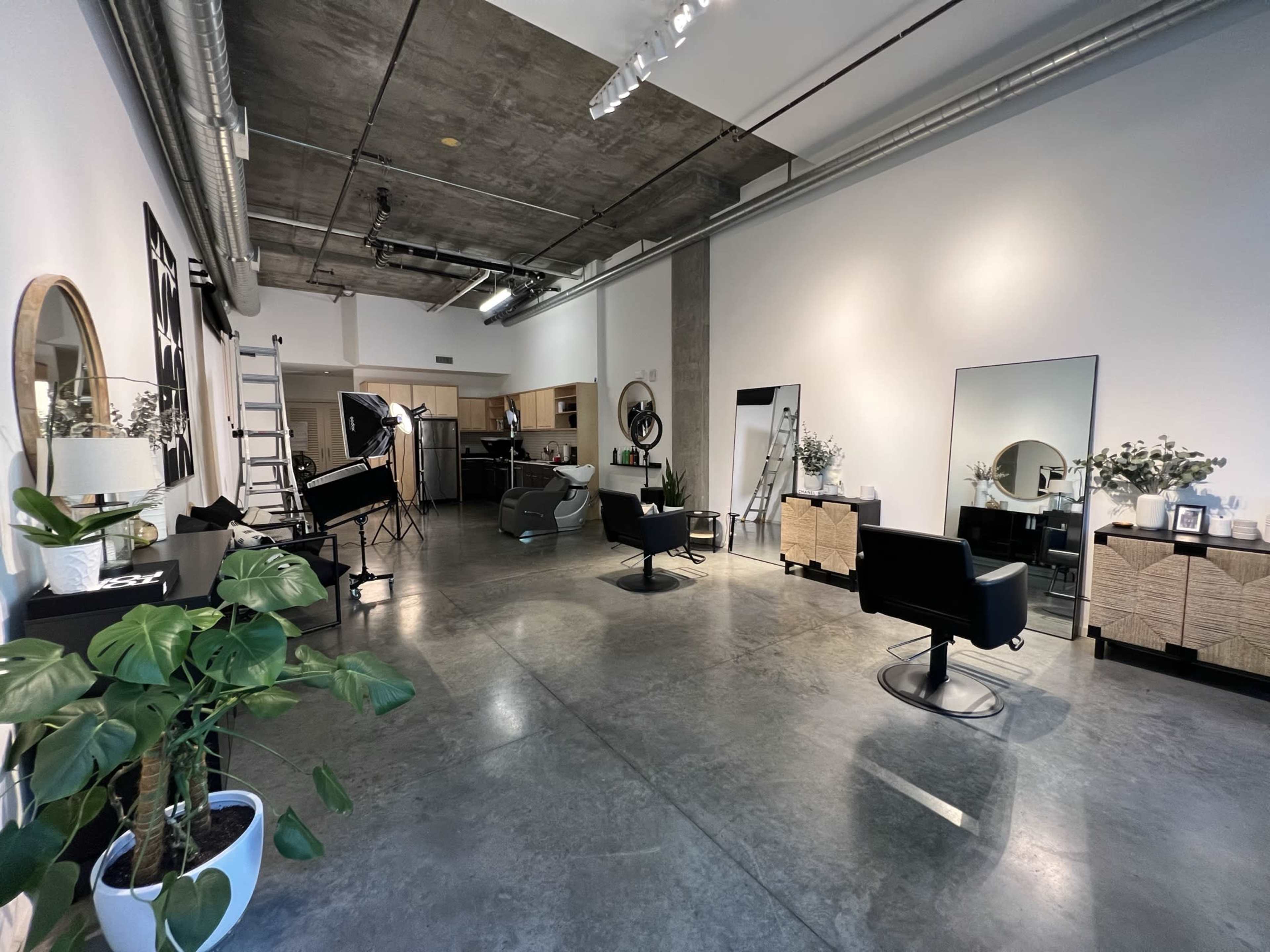 A spacious, modern hair salon with several styling chairs, mirrors, and a few plants, featuring concrete floors and industrial-style ceilings.