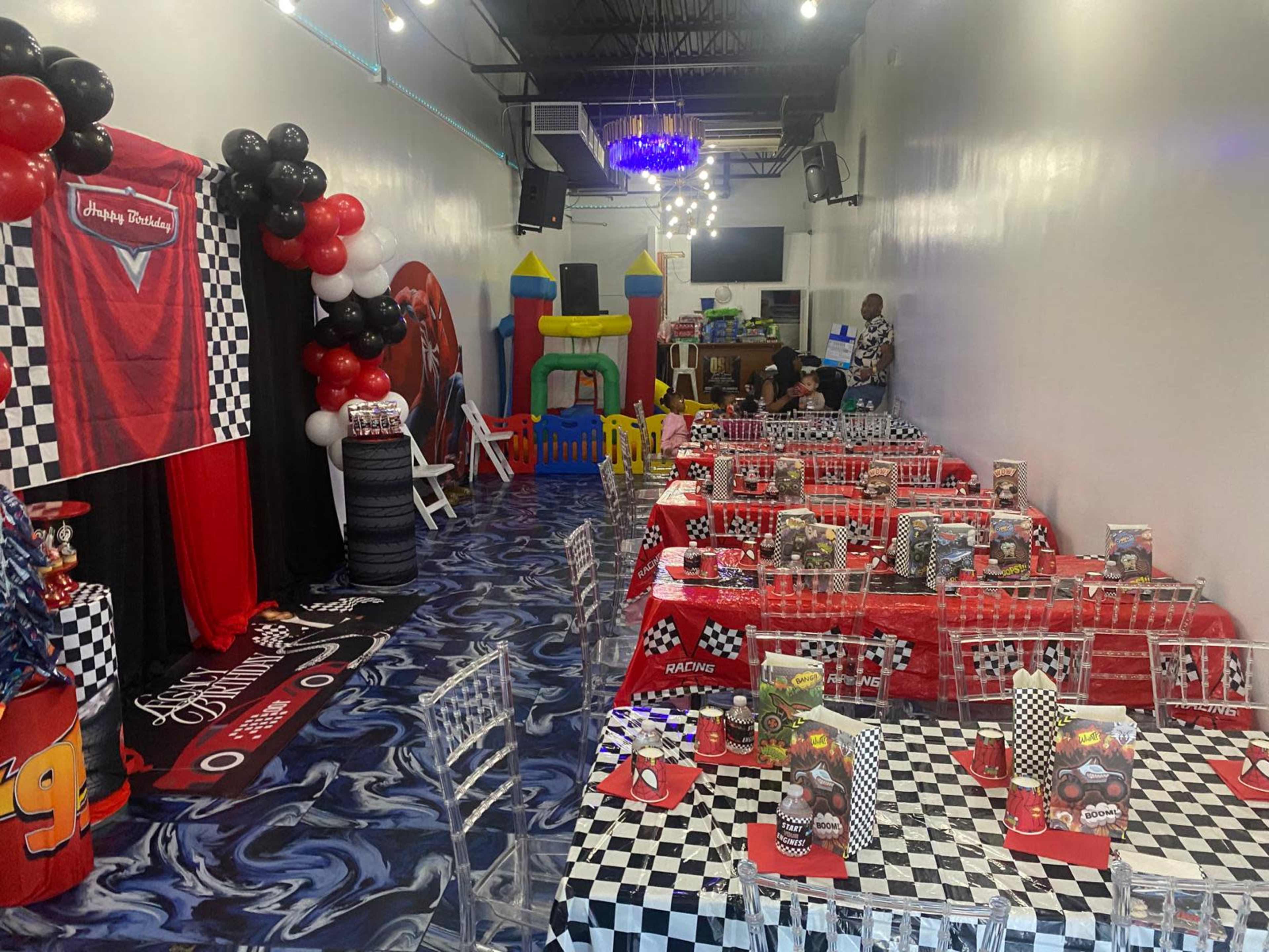 The image shows a decorated party venue with several tables set up in a racing theme, featuring red and black tablecloths, checkered decor, and a bouncy castle in the background.