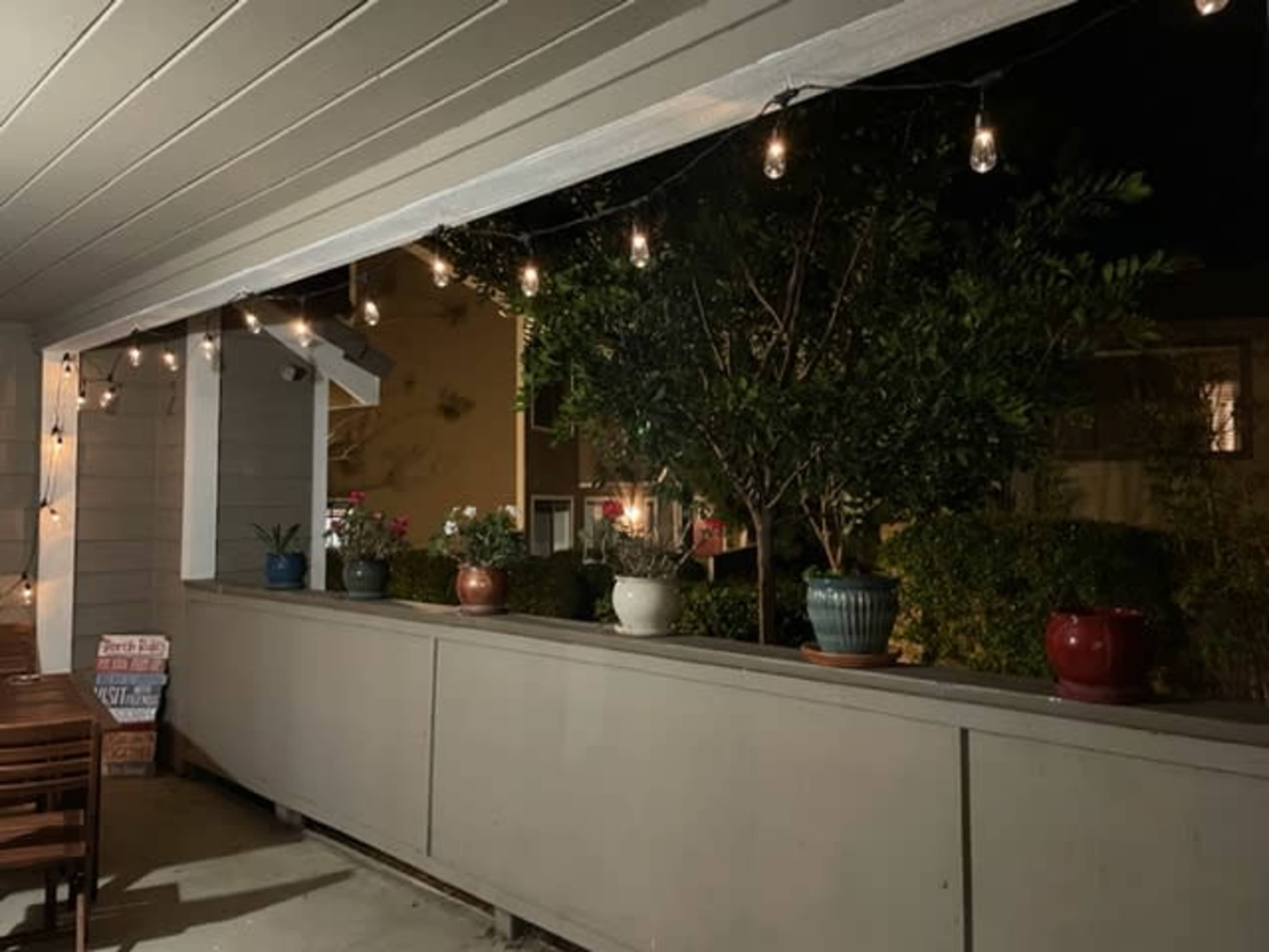A porch area is illuminated by string lights, featuring potted plants along a railing.
