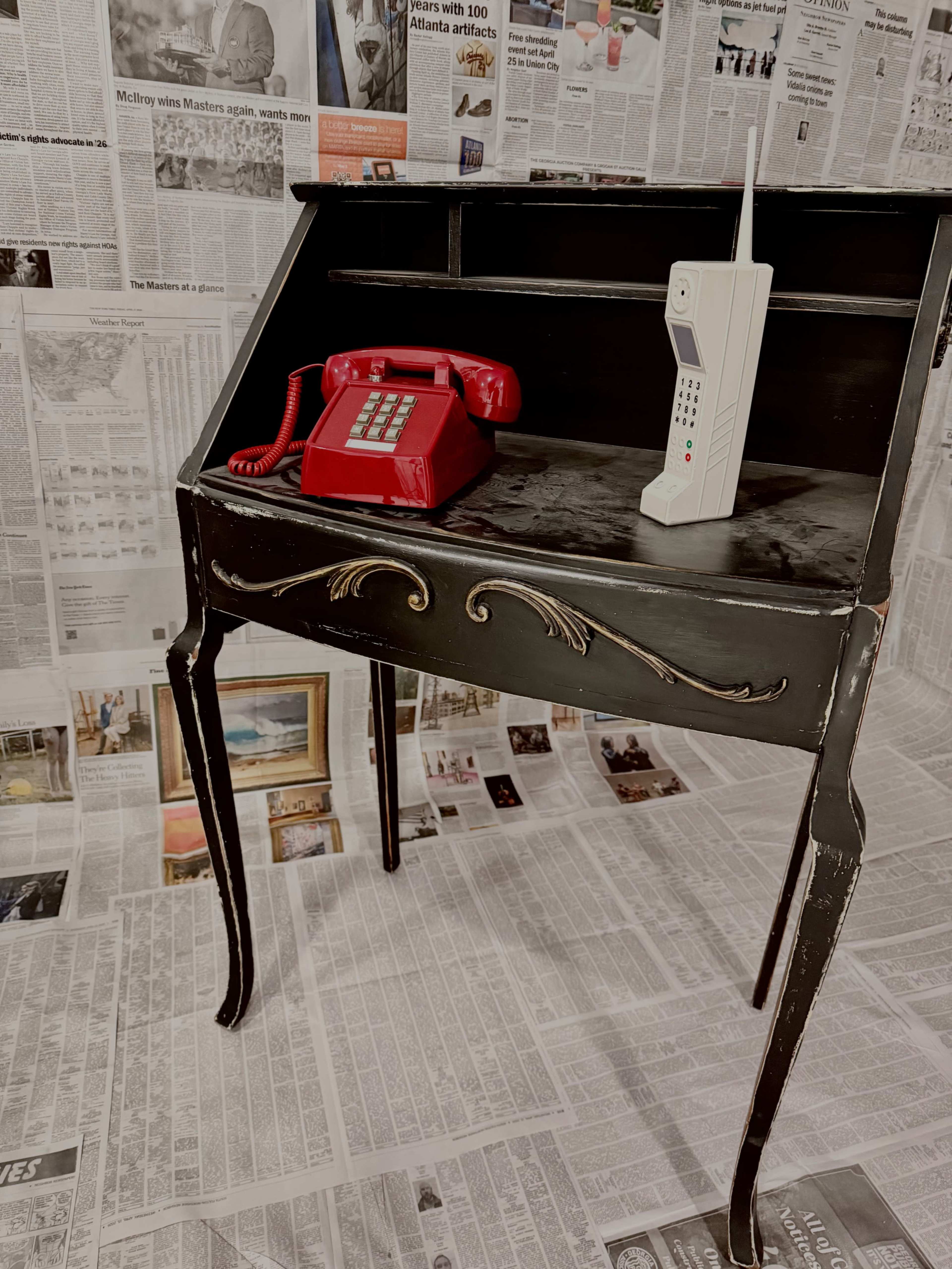 A black writing desk with ornate legs displays a red rotary phone and a white cordless phone against a backdrop of newspaper clippings.