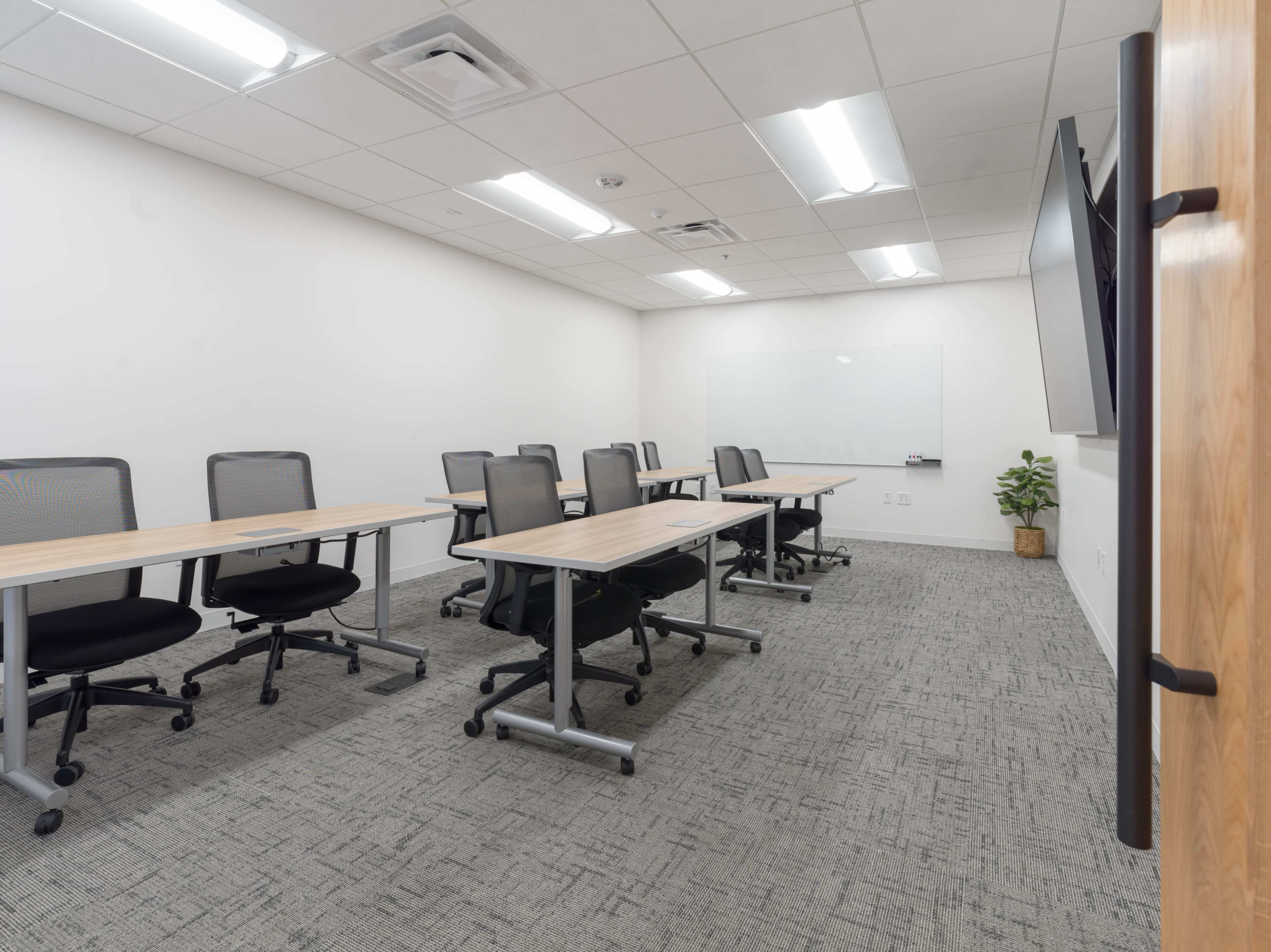 A modern meeting room features several rows of tables and chairs, with a whiteboard and a large screen at one end.