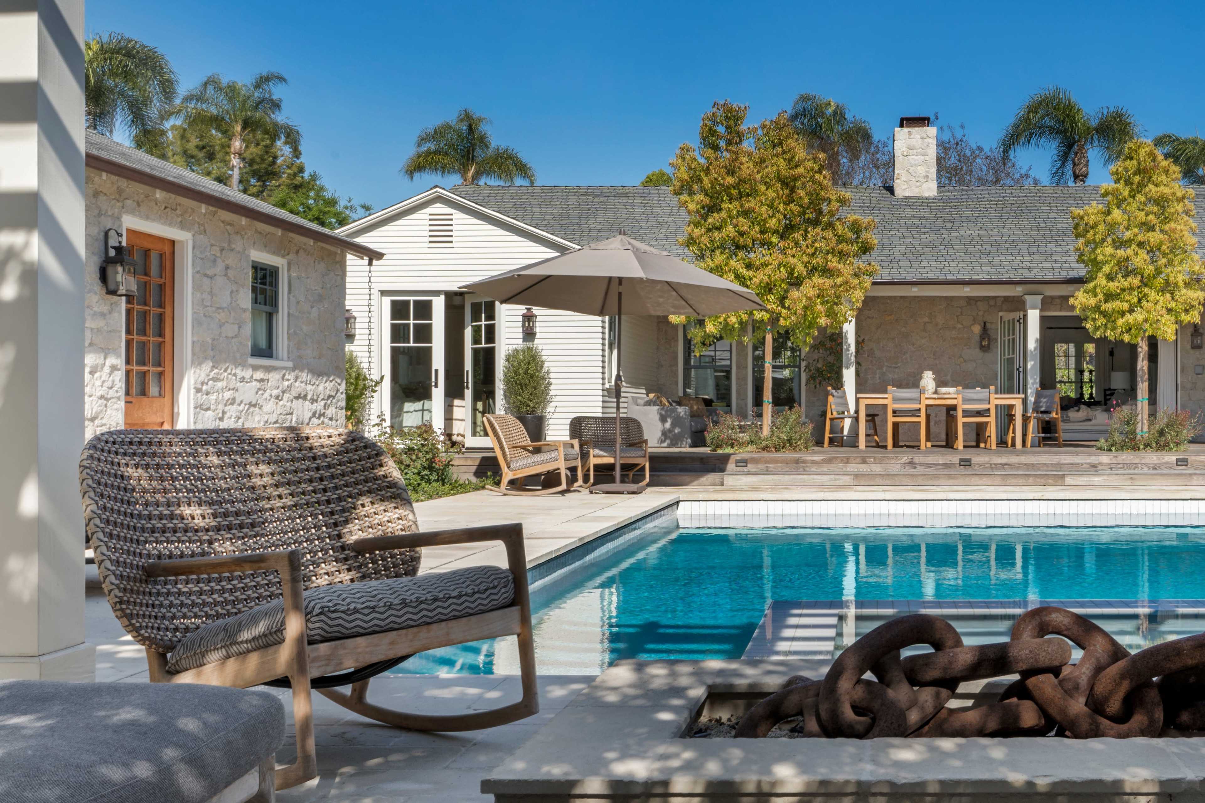 A modern backyard features a swimming pool with lounge chairs, an umbrella, and a dining area surrounded by greenery and stone architecture.