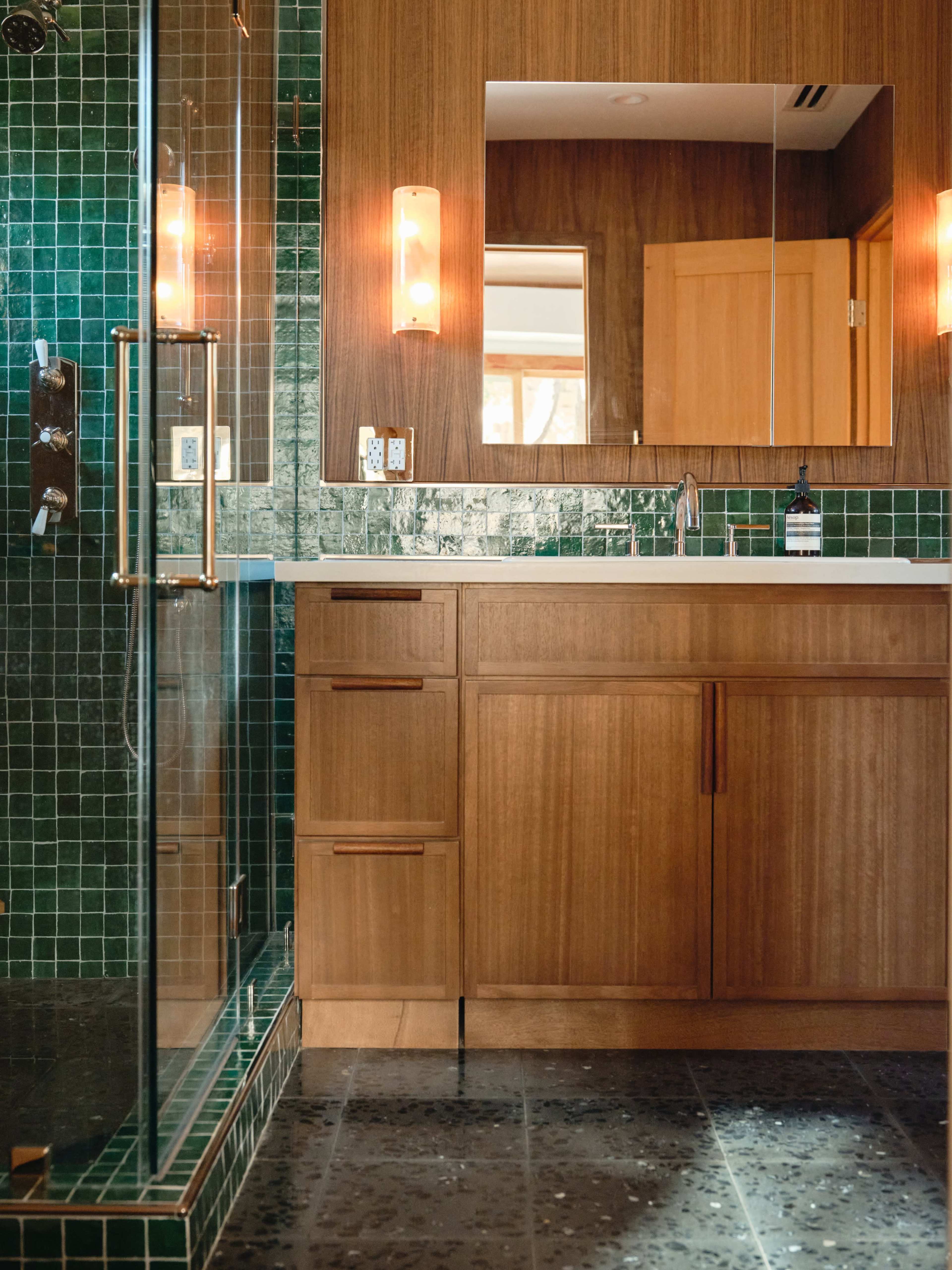 The image shows a modern bathroom featuring a glass shower, wooden cabinetry, and green tiled walls.