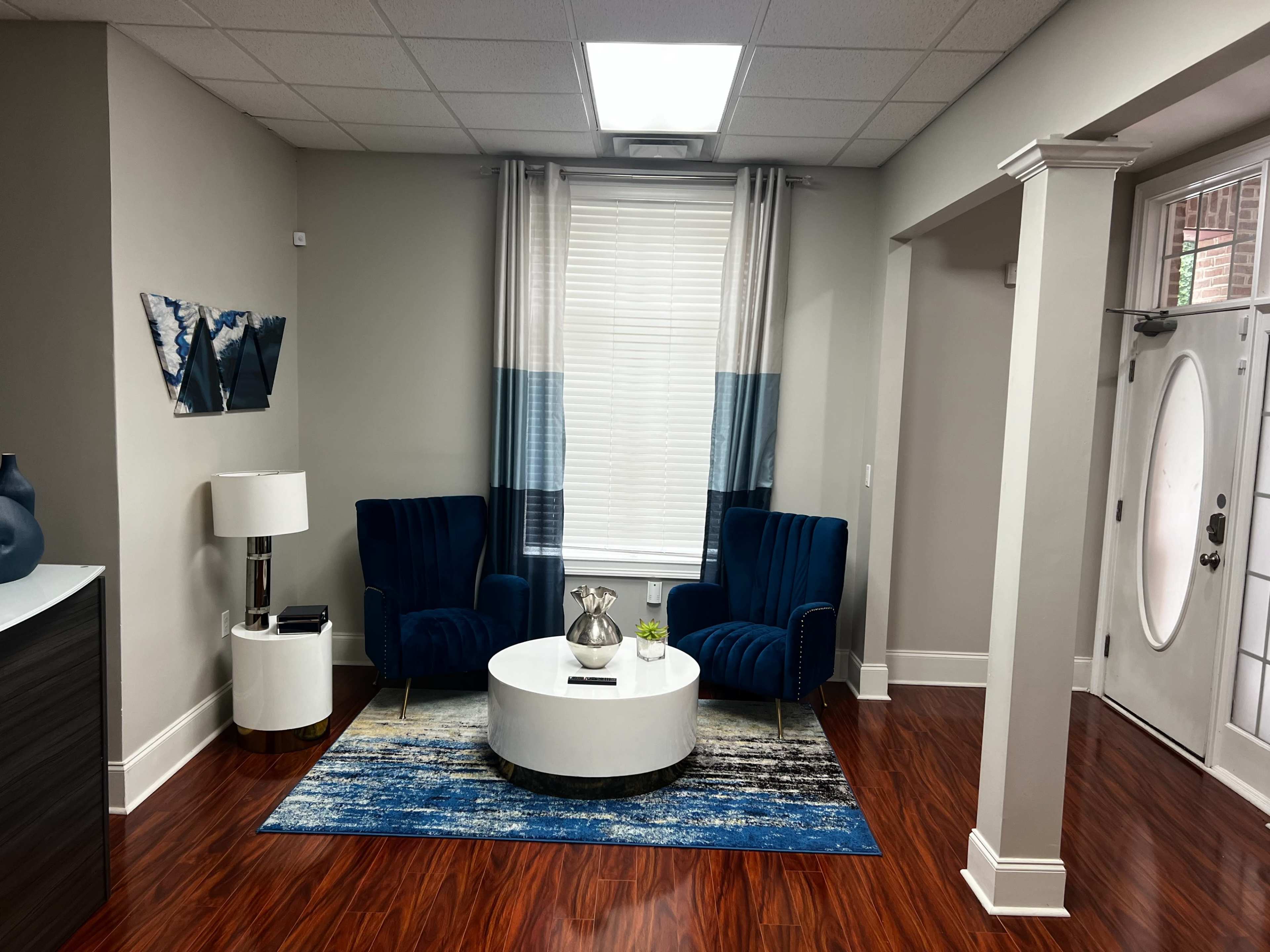 The image shows a modern sitting area featuring two blue velvet chairs, a round white table, and a large area rug, with a front door visible in the background.