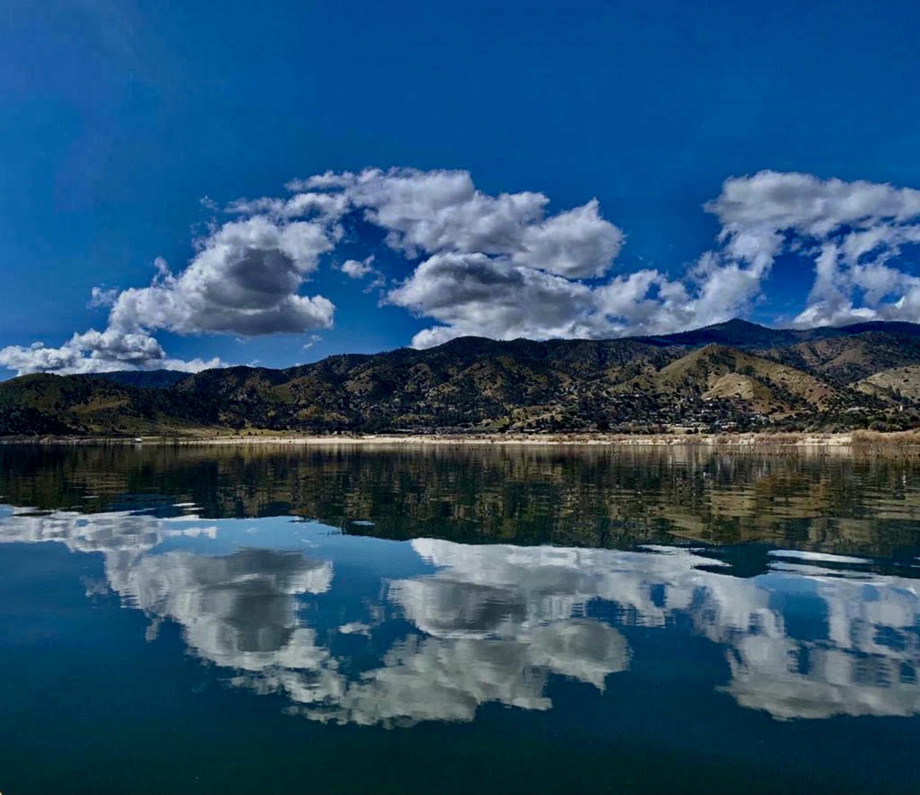 A calm lake bordered by mountains, with fluffy clouds reflected on the water's surface.