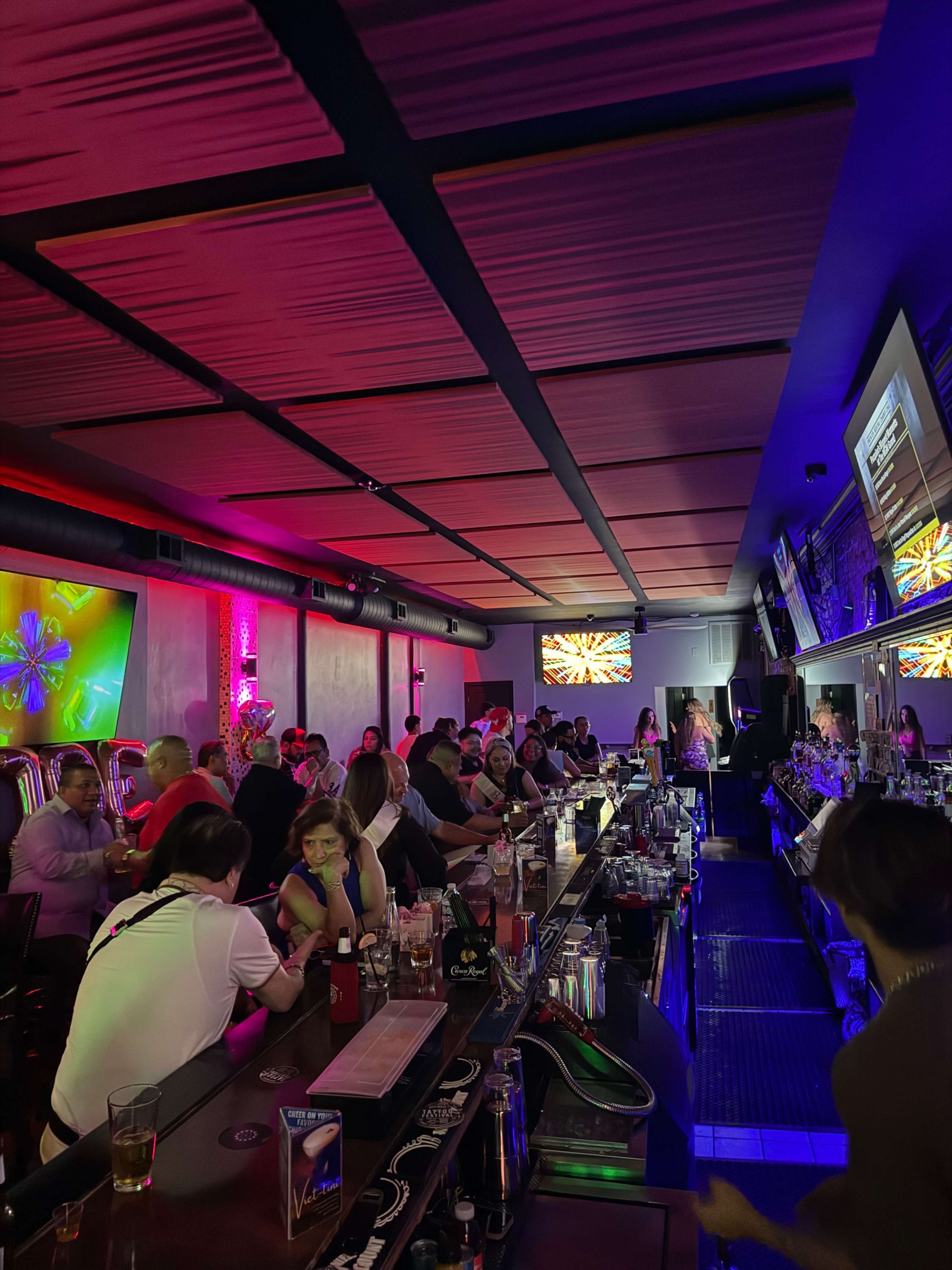A lively bar scene features patrons seated at a long counter and tables, with colorful lighting and multiple screens displaying vibrant visuals.