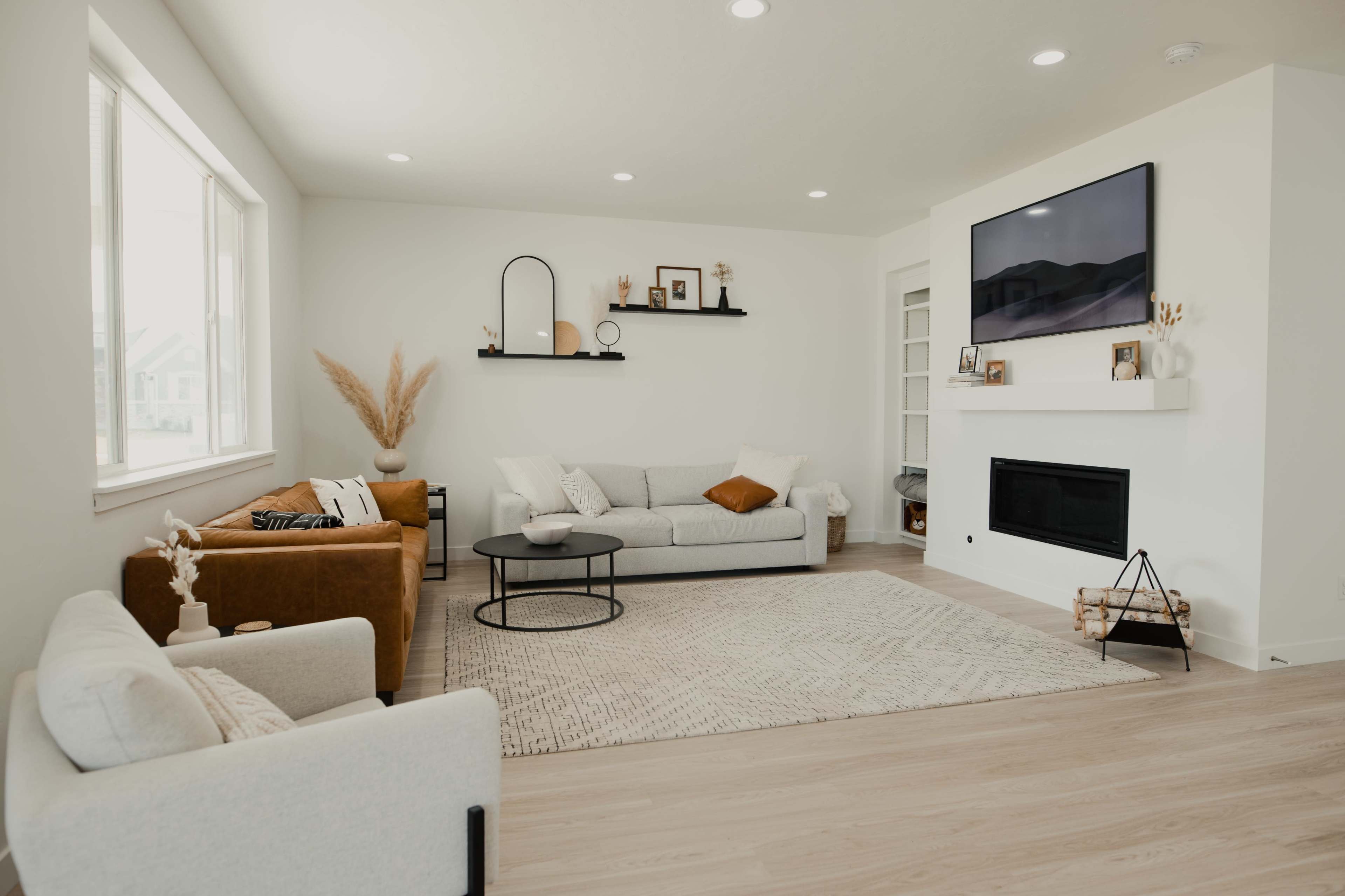 The room features a modern living space with a gray sectional sofa, a brown armchair, a circular coffee table, and a large window allowing natural light to enter.