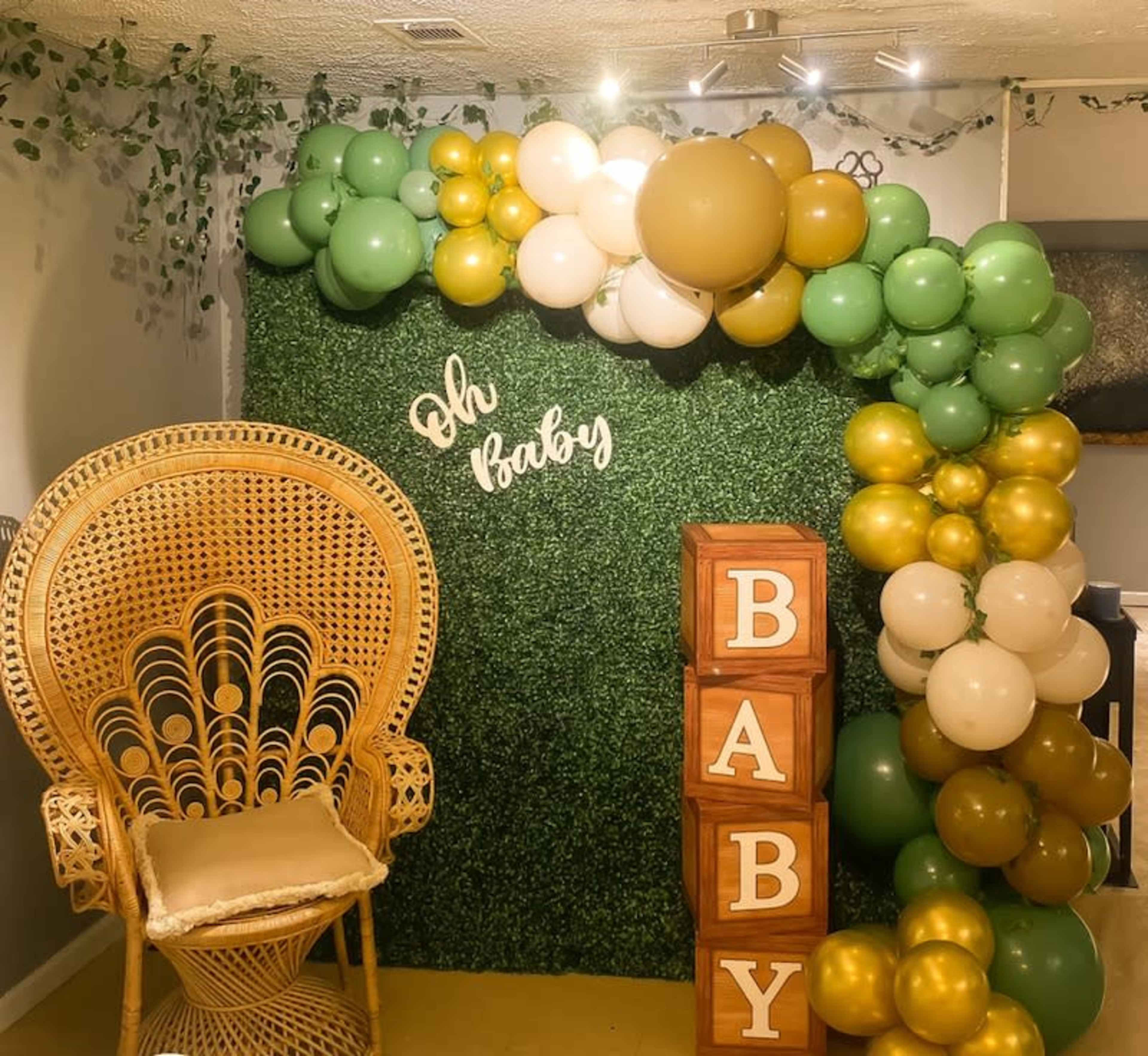The image shows a decorated wall with green foliage, a balloon arrangement in green, white, and gold, and stacked wooden blocks spelling "BABY," next to a large wicker chair.