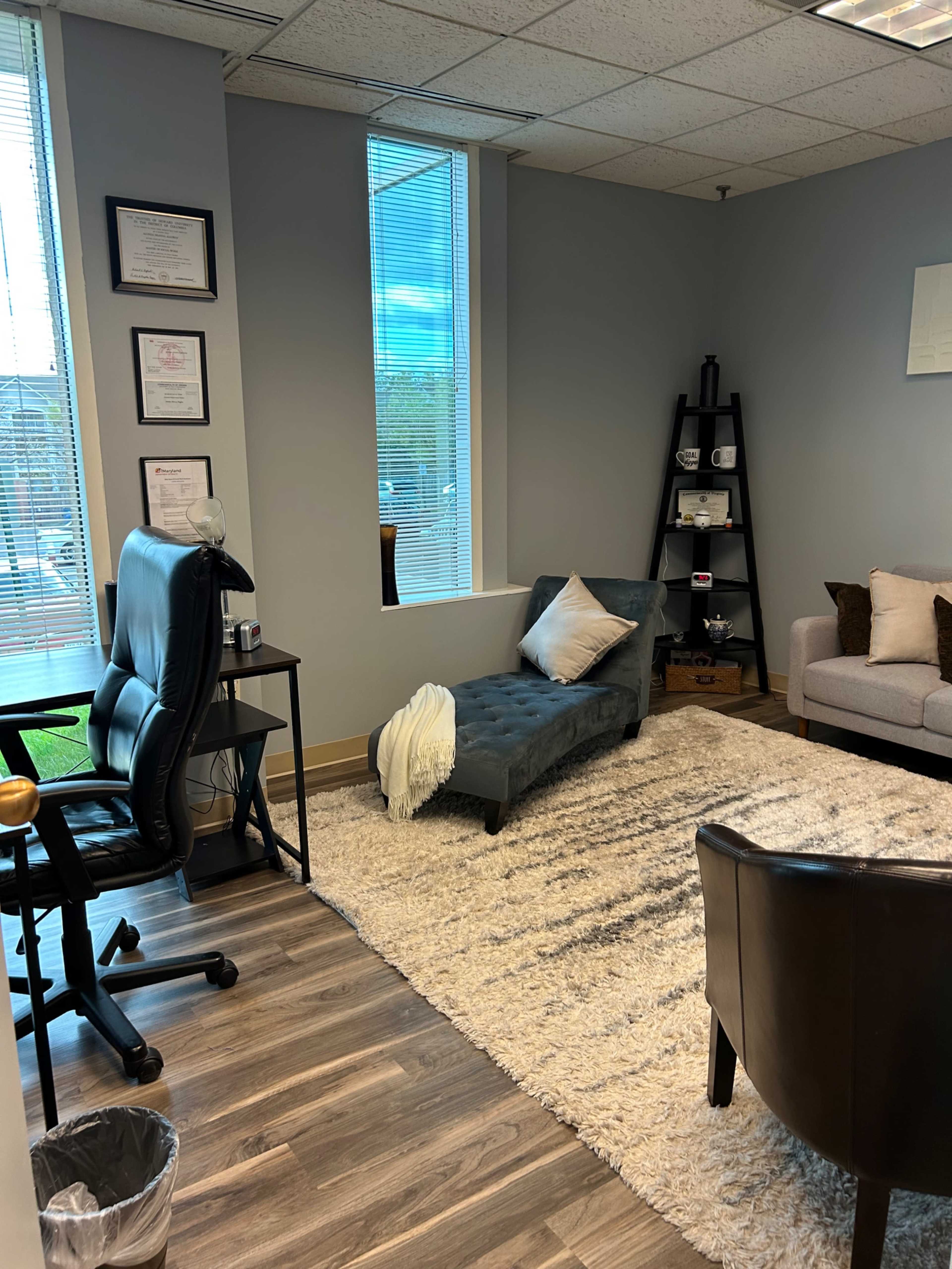 The image shows a modern office space with a cozy seating area, including a chaise lounge, a brown chair, and a small shelf with decorative items.