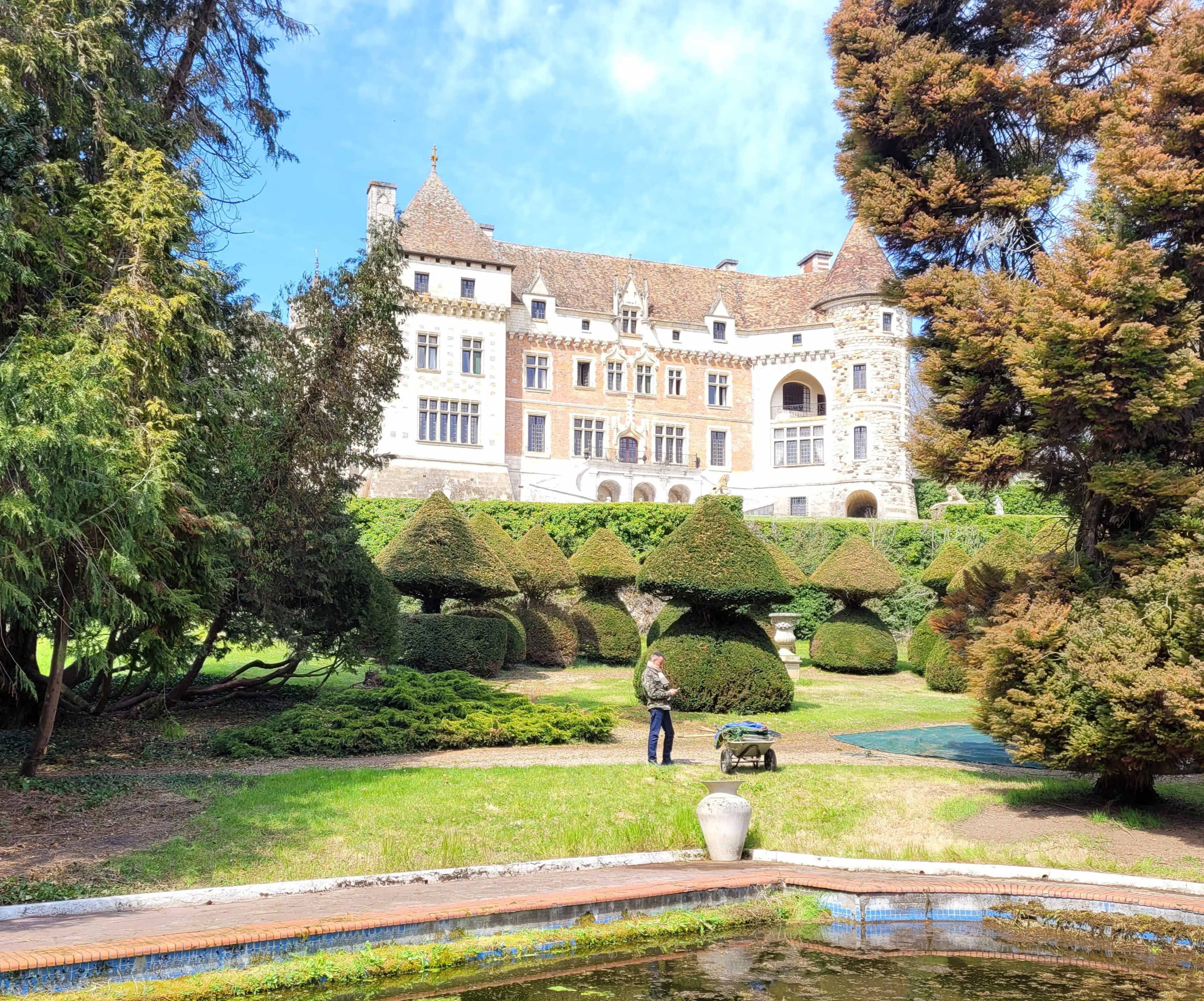 A gardener works among neatly trimmed hedges in front of a large stone castle surrounded by lush greenery.