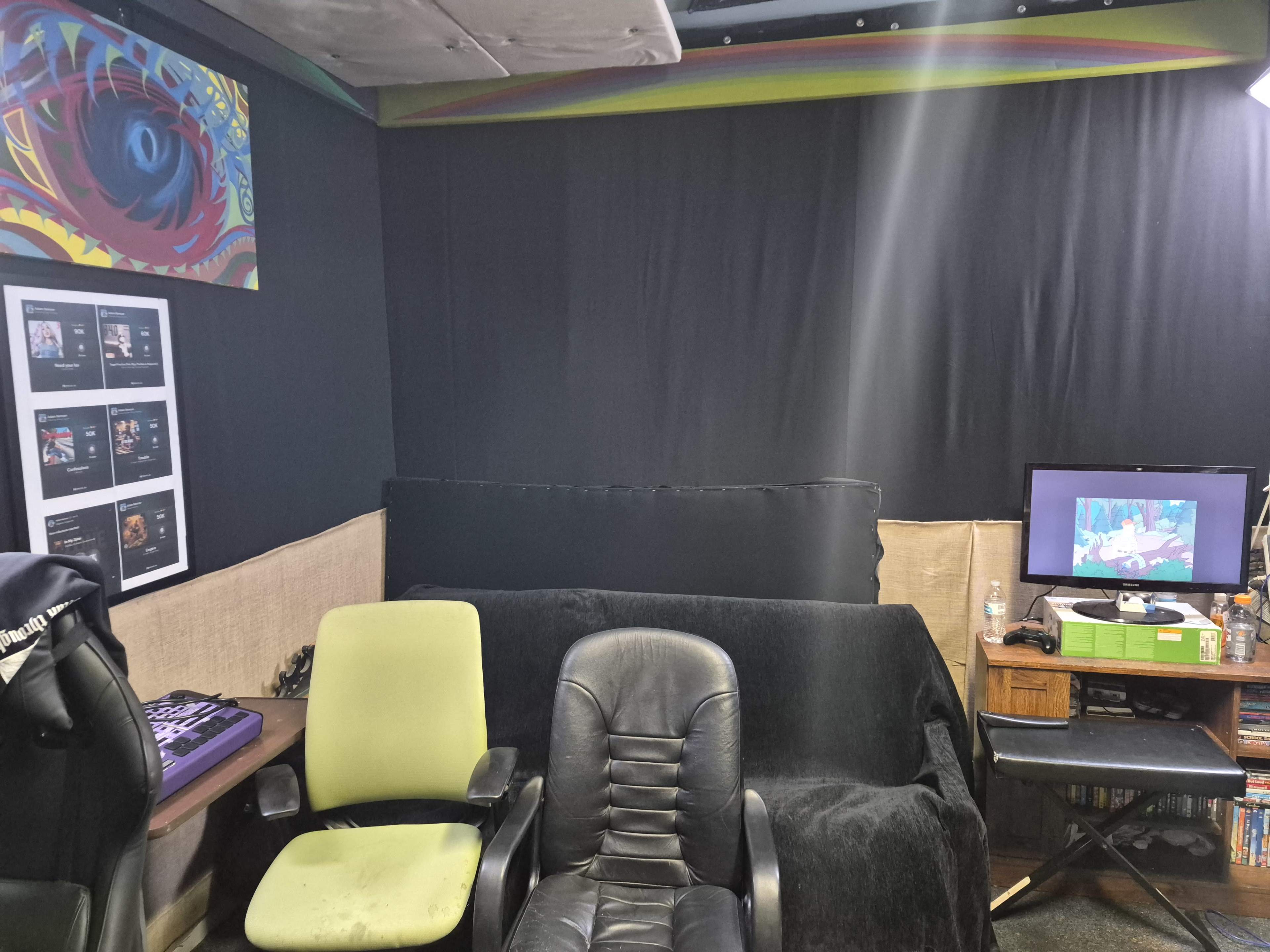 The image shows a small room with two office chairs, a couch covered with a black blanket, a television displaying animated content, and framed posters on the wall.