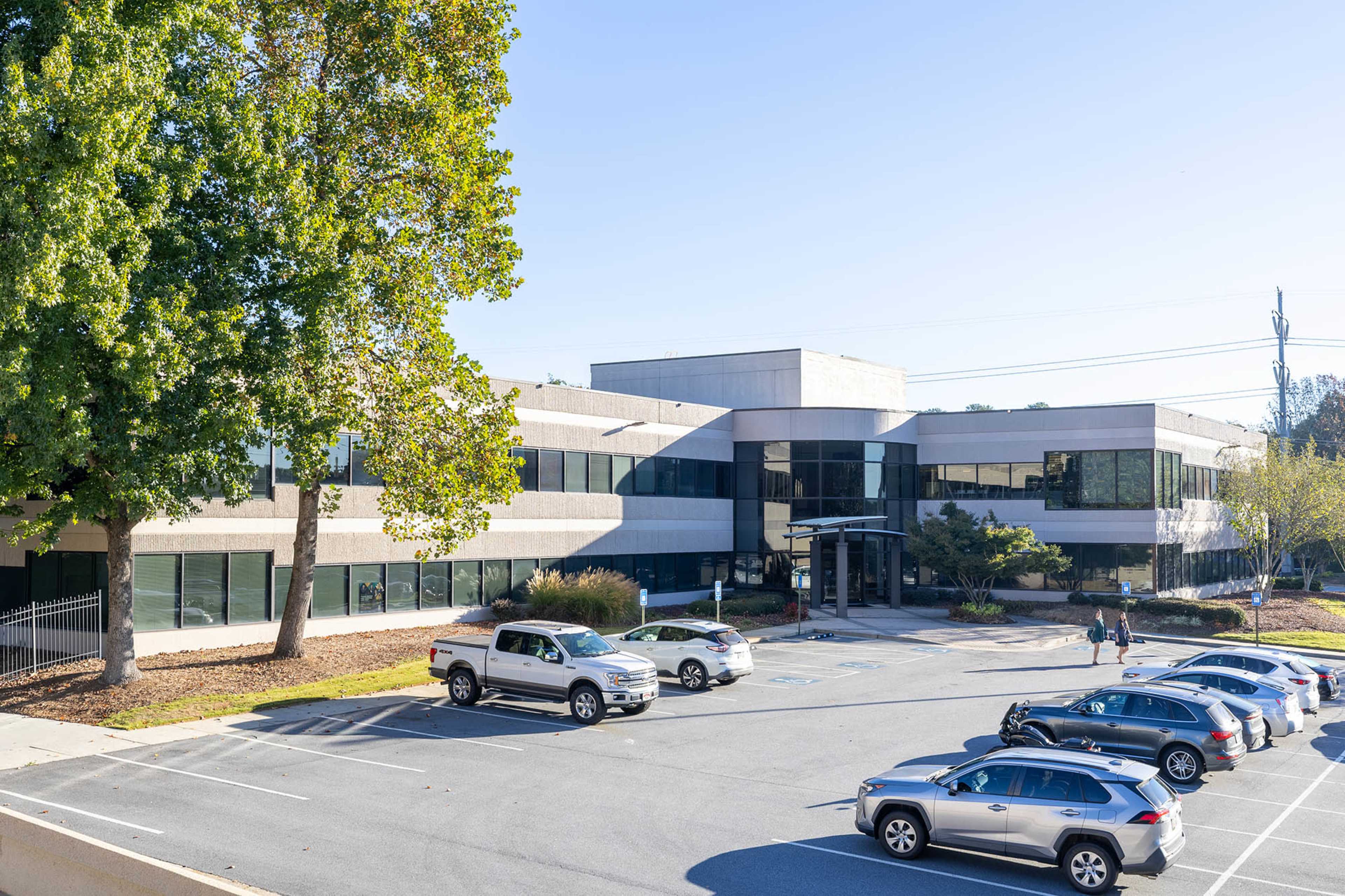 A modern, two-story office building with large windows and a parking lot filled with vehicles is depicted.