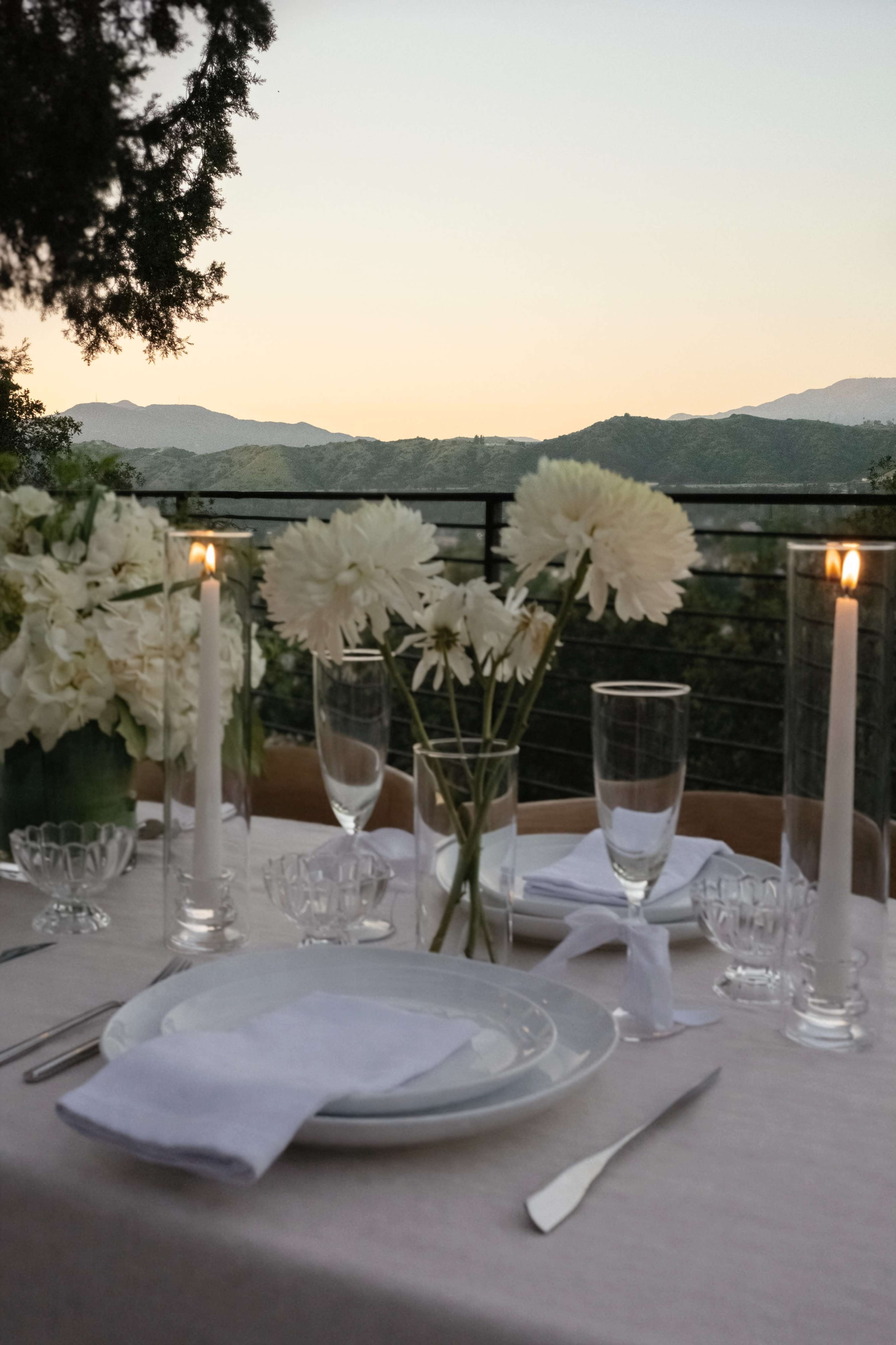 A dining table is set with white flowers, glassware, and candles, overlooking a scenic mountain view at sunset.