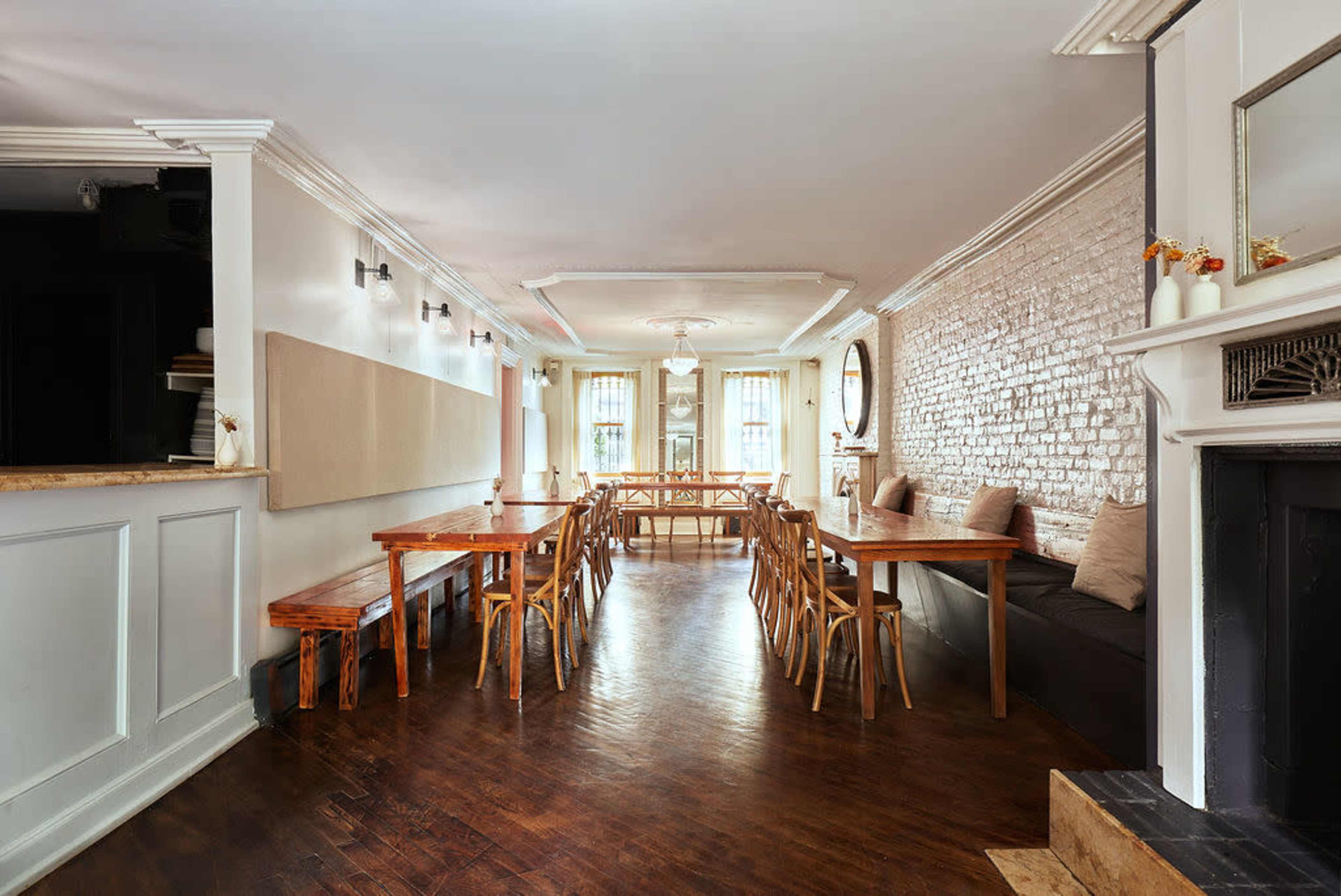 The image shows a spacious dining area featuring wooden tables and chairs, with a light-colored brick wall and large windows allowing natural light to fill the room.