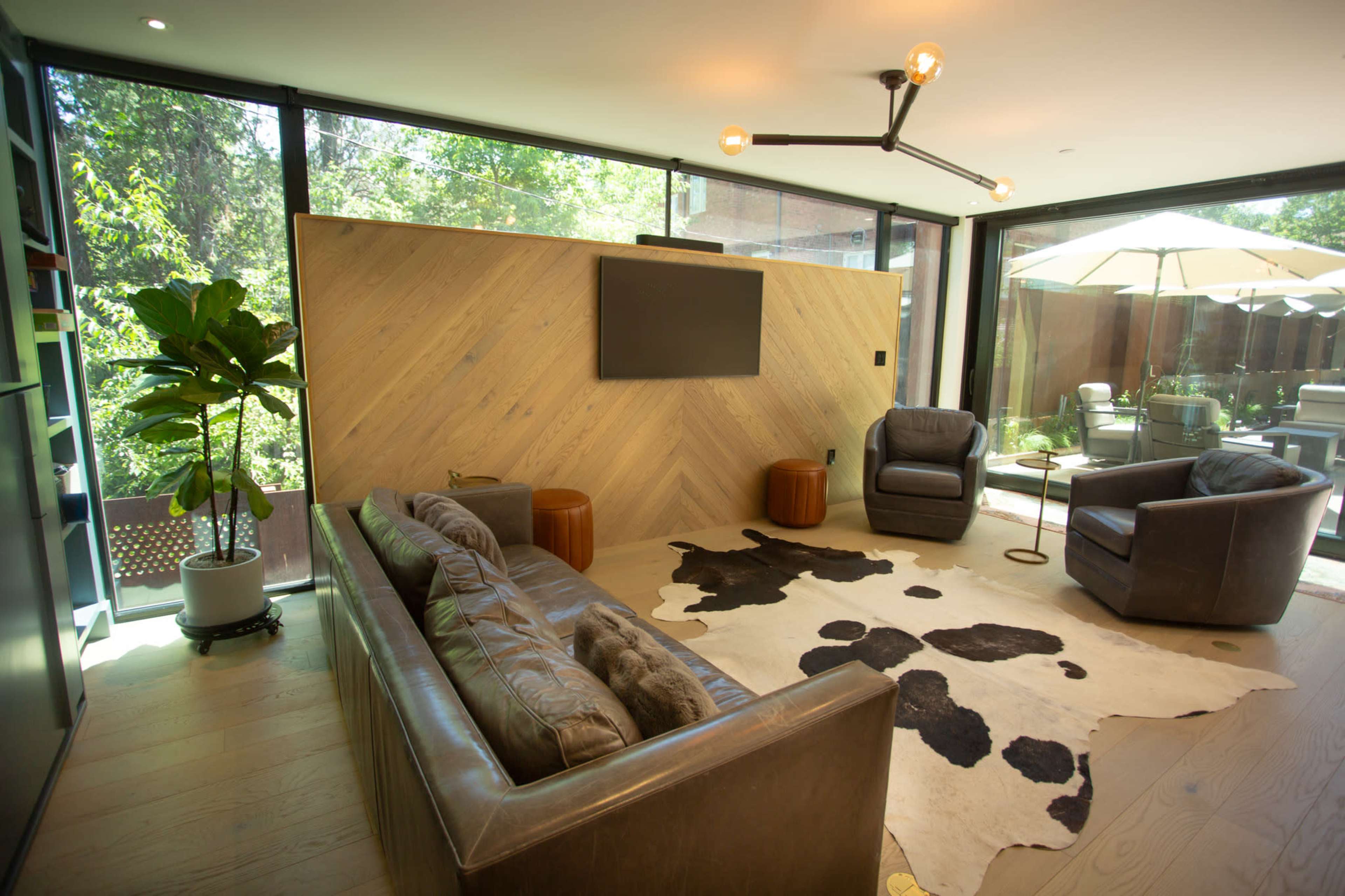 The image shows a modern living room featuring a leather sofa, two armchairs, a cowhide rug, a wooden accent wall, and large windows that open to a patio with umbrellas.