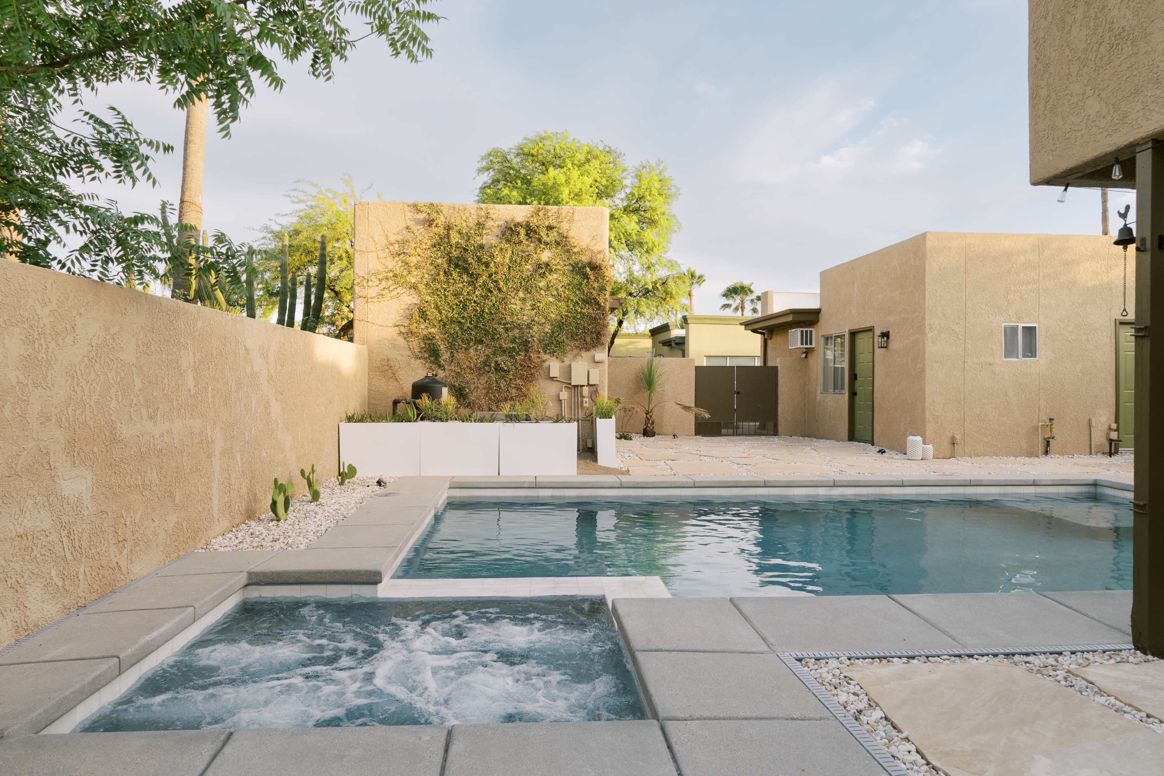 A modern backyard with a swimming pool and spa, surrounded by beige stucco walls and landscaped areas.
