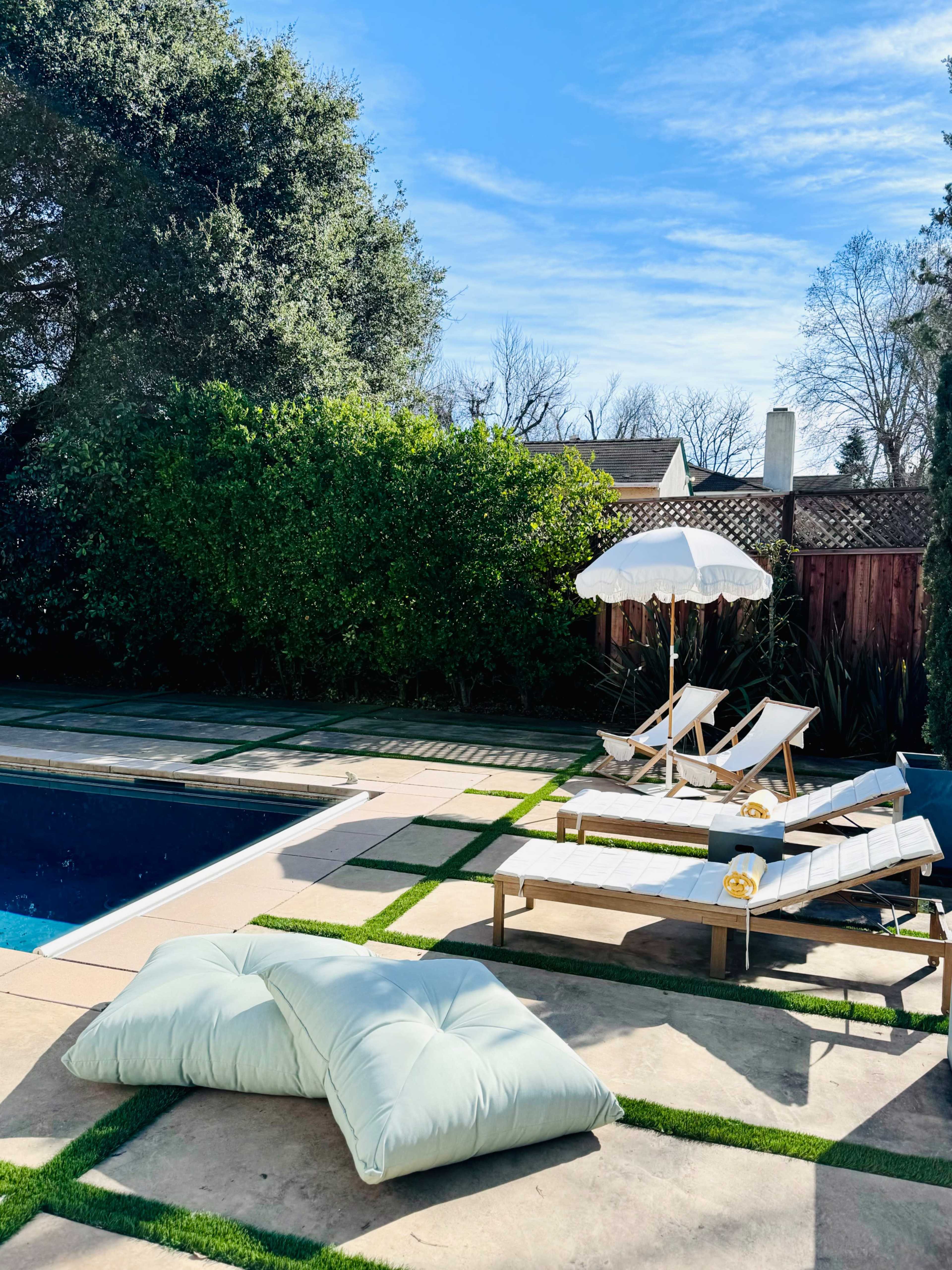 A backyard pool area with lounge chairs, an umbrella, and oversized cushions arranged on the patio beside the water.