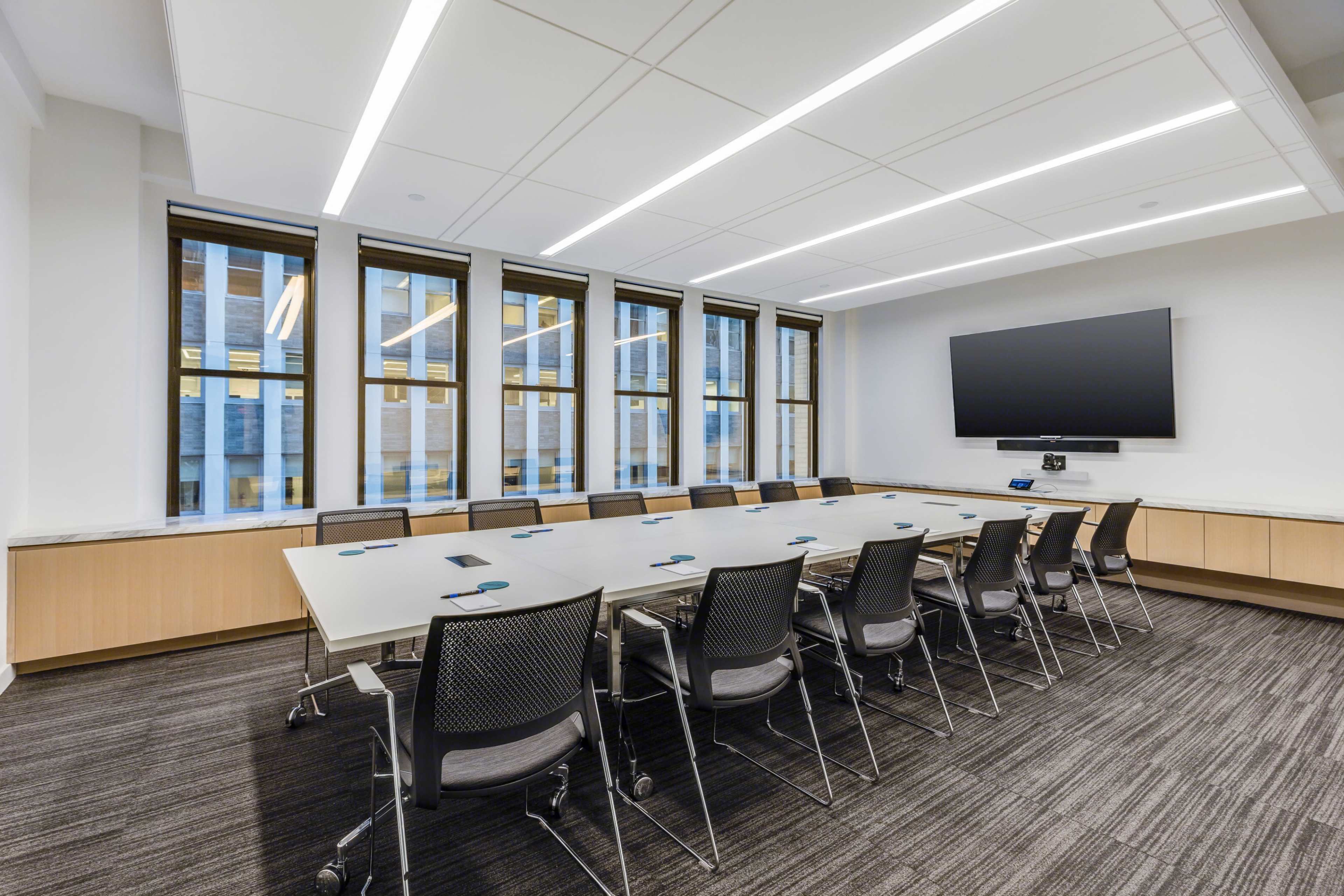 A modern conference room features a long table with ergonomic chairs, large windows allowing natural light, and a wall-mounted television.