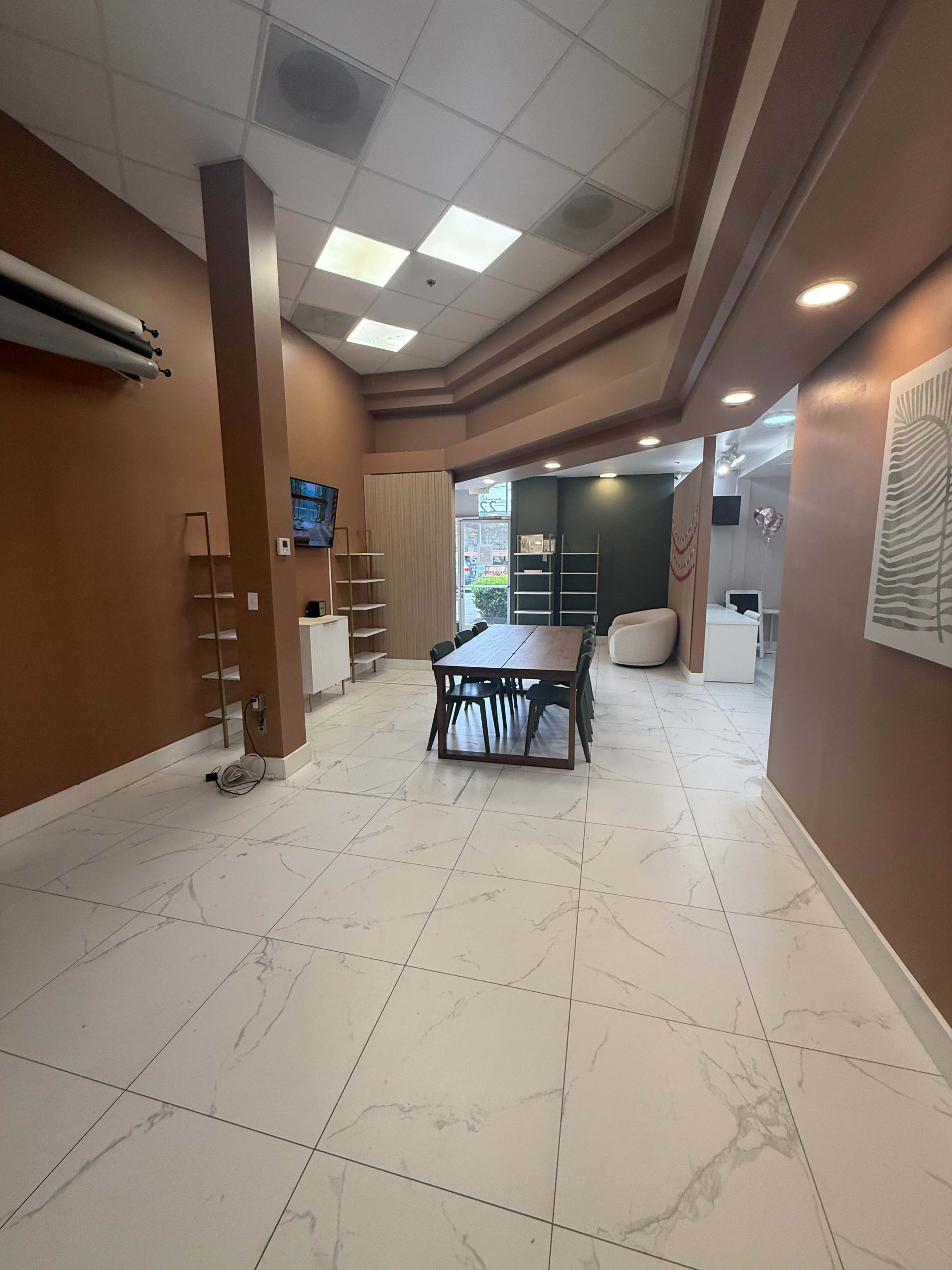 The image shows a spacious, well-lit interior of a modern office or reception area with a large table in the center and shelves along the walls.