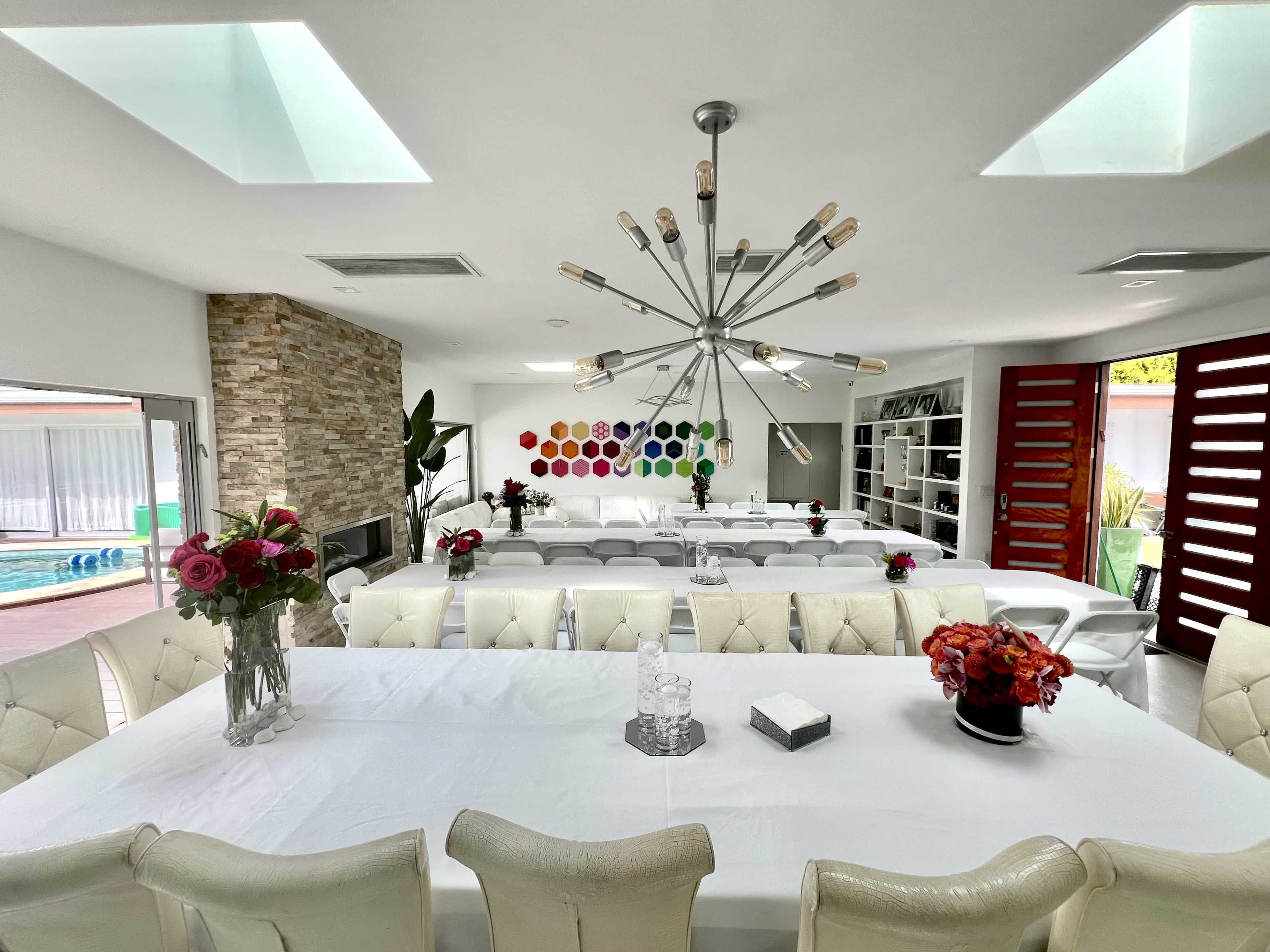 A modern dining area featuring a long white table surrounded by tufted chairs, a stone accent wall, and a colorful art installation hanging above the table.