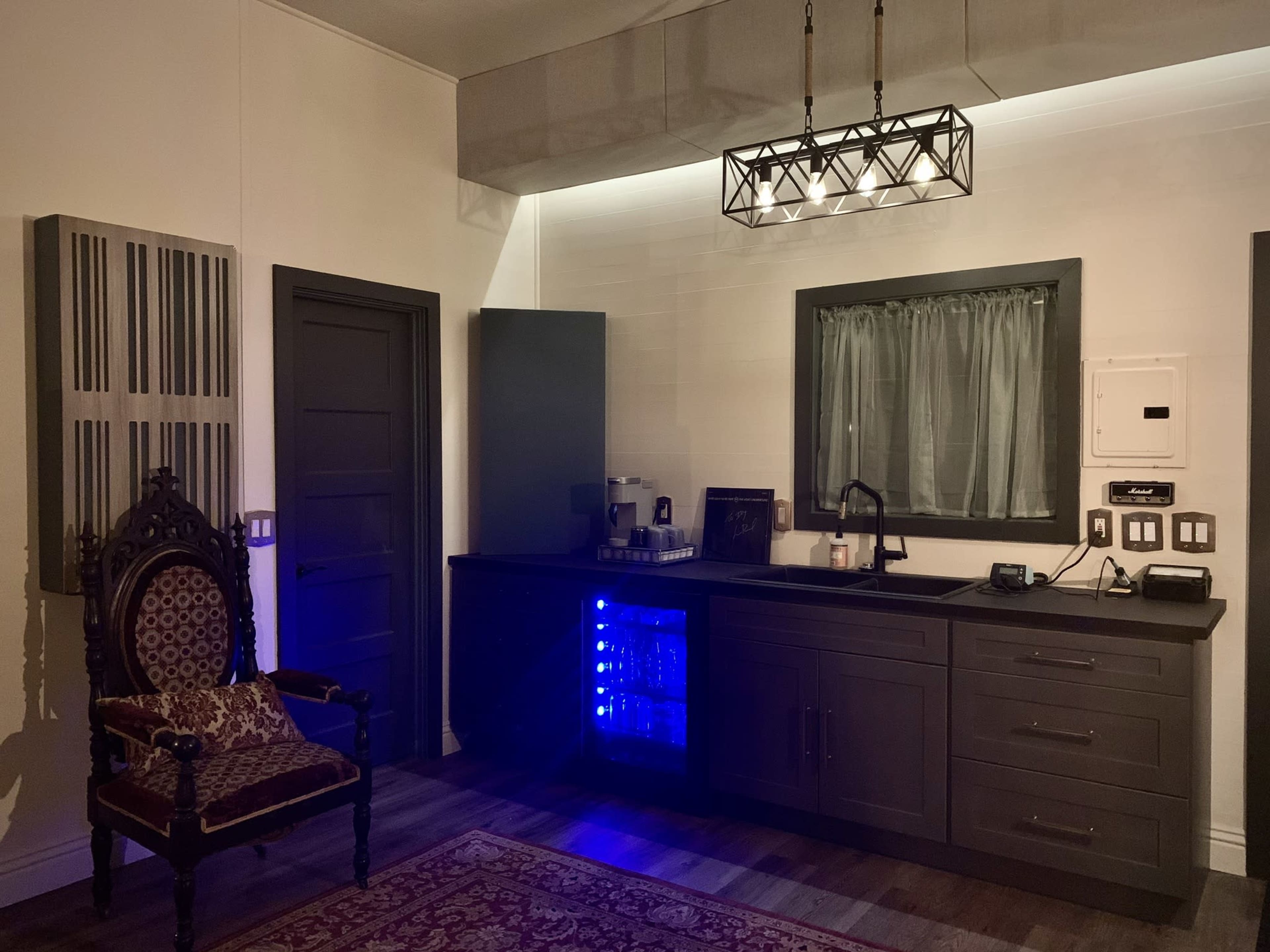 A kitchen area featuring a dark cabinet with a blue-lit mini refrigerator, a sink, and a vintage-style chair beside it.