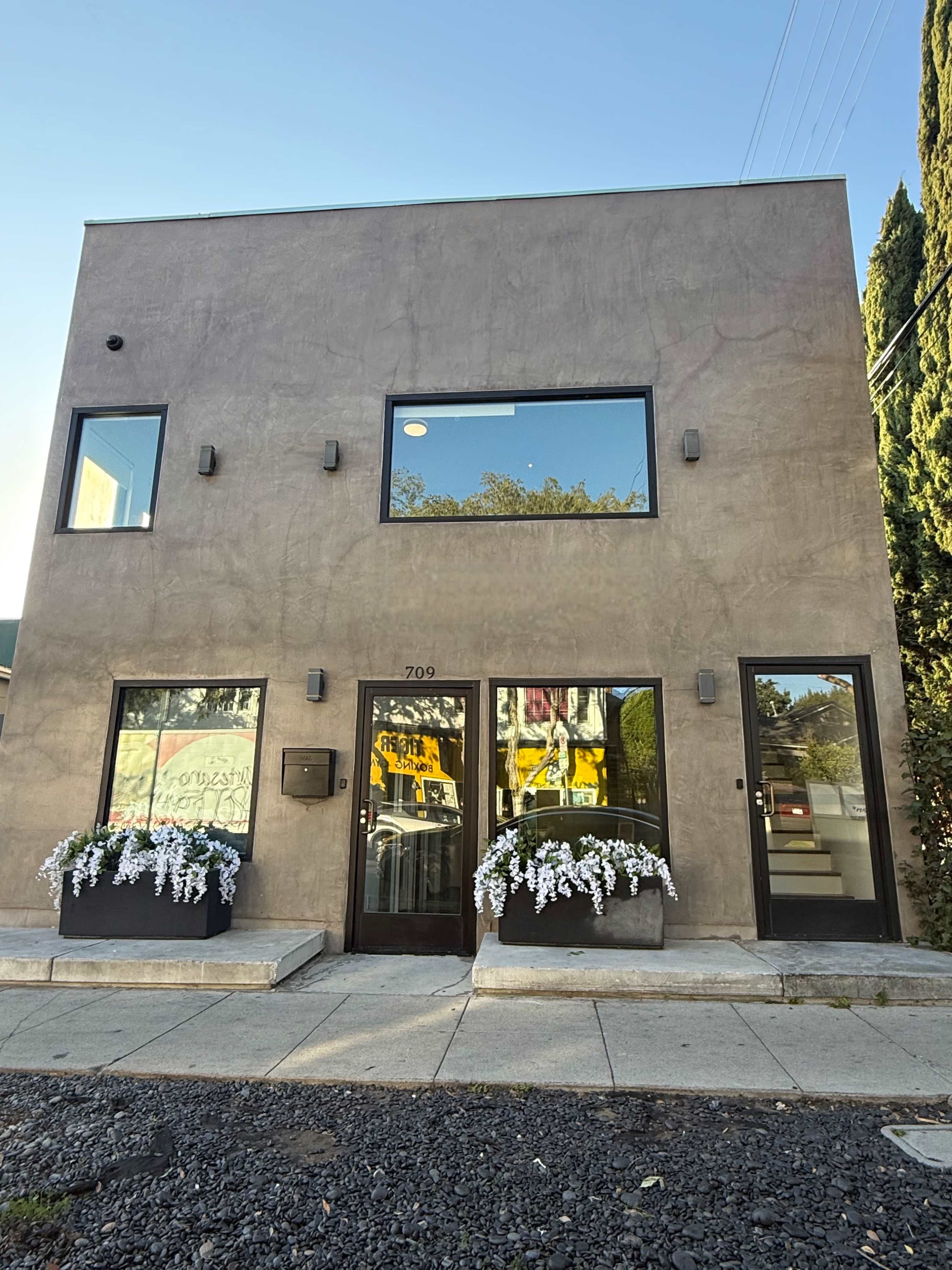 The image shows a modern two-story building with large windows and planters filled with white flowers at the entrance.