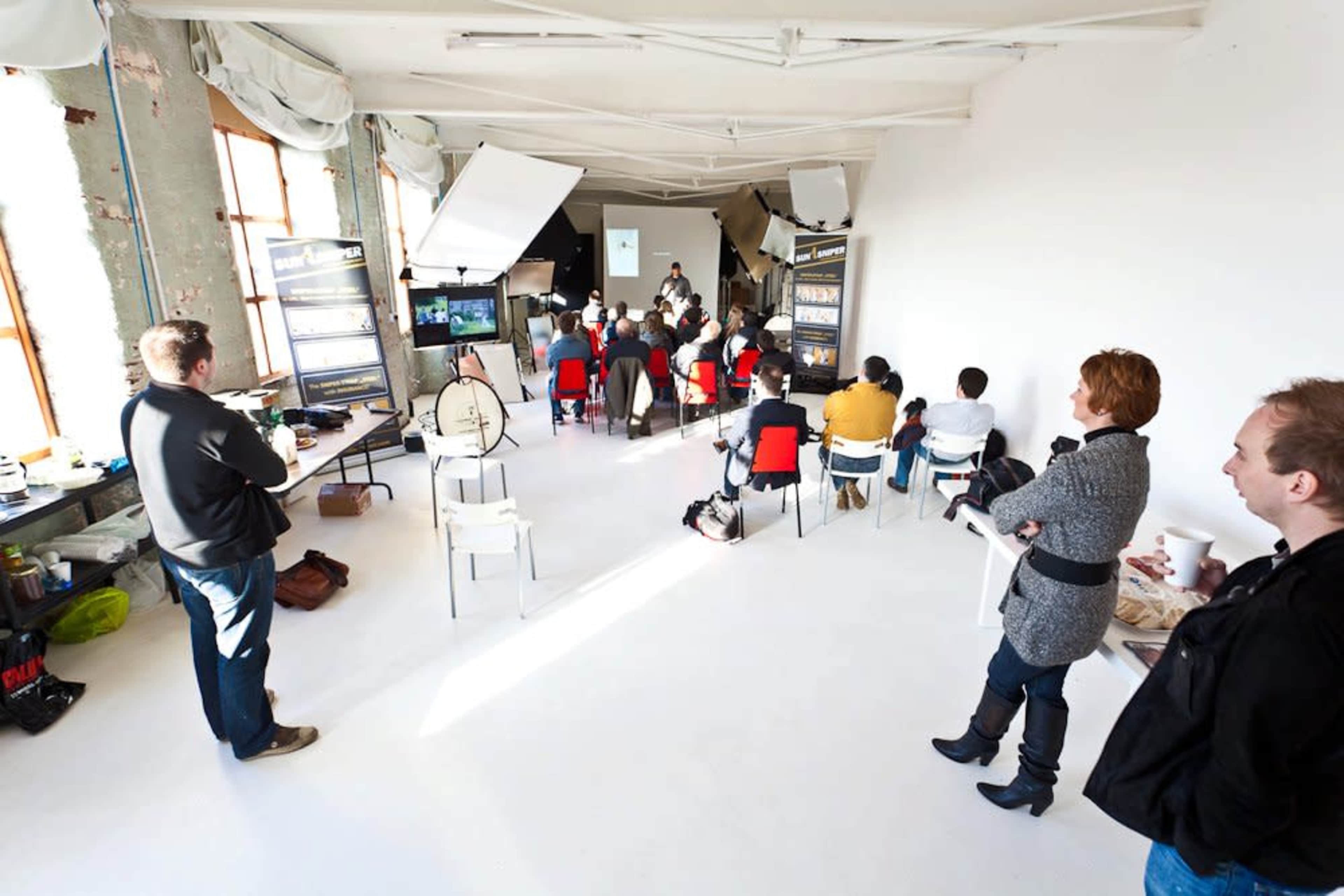 A group of people sits in chairs facing a presentation at the front of a well-lit, spacious room with minimal decor.