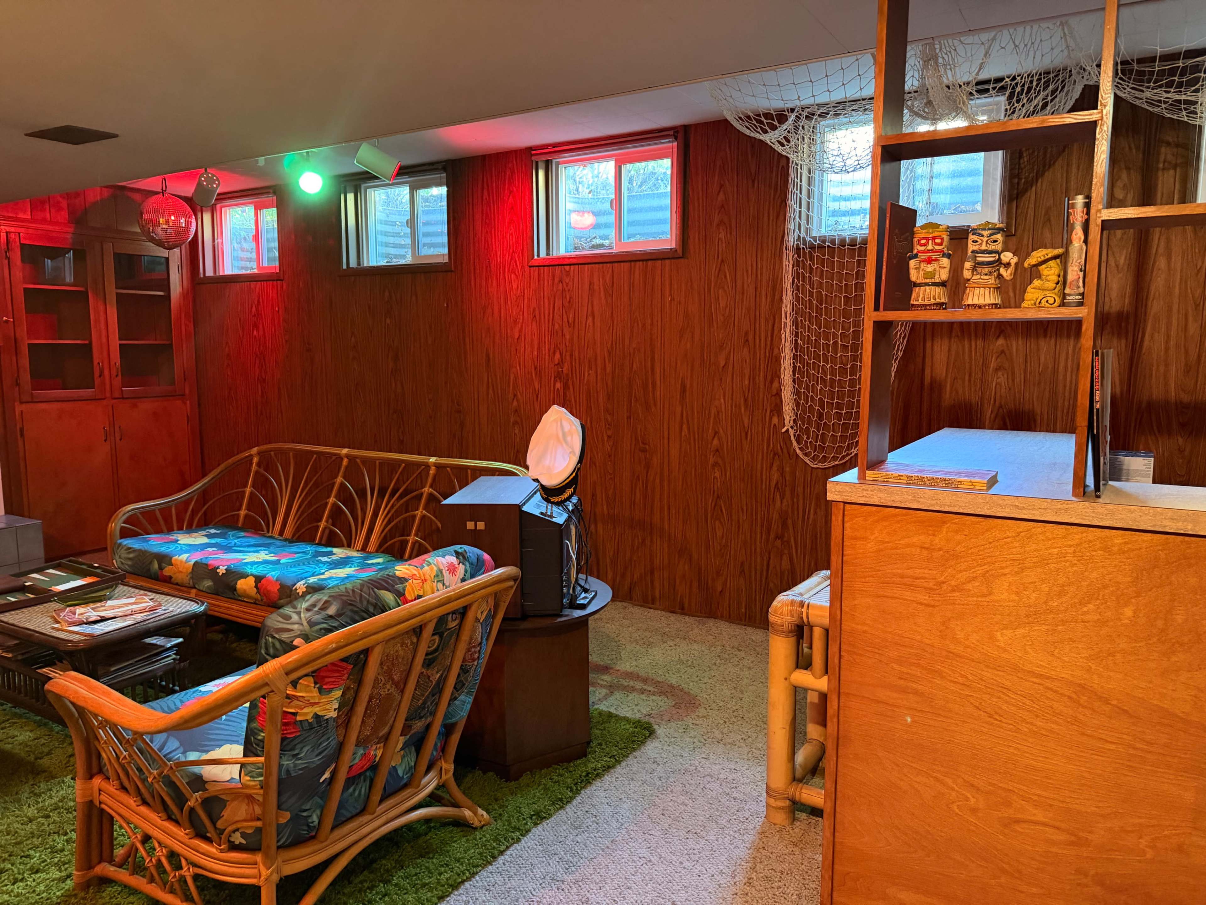 The image shows a basement living area with a colorful couch, a wooden bookshelf, and light fixtures casting a warm glow.