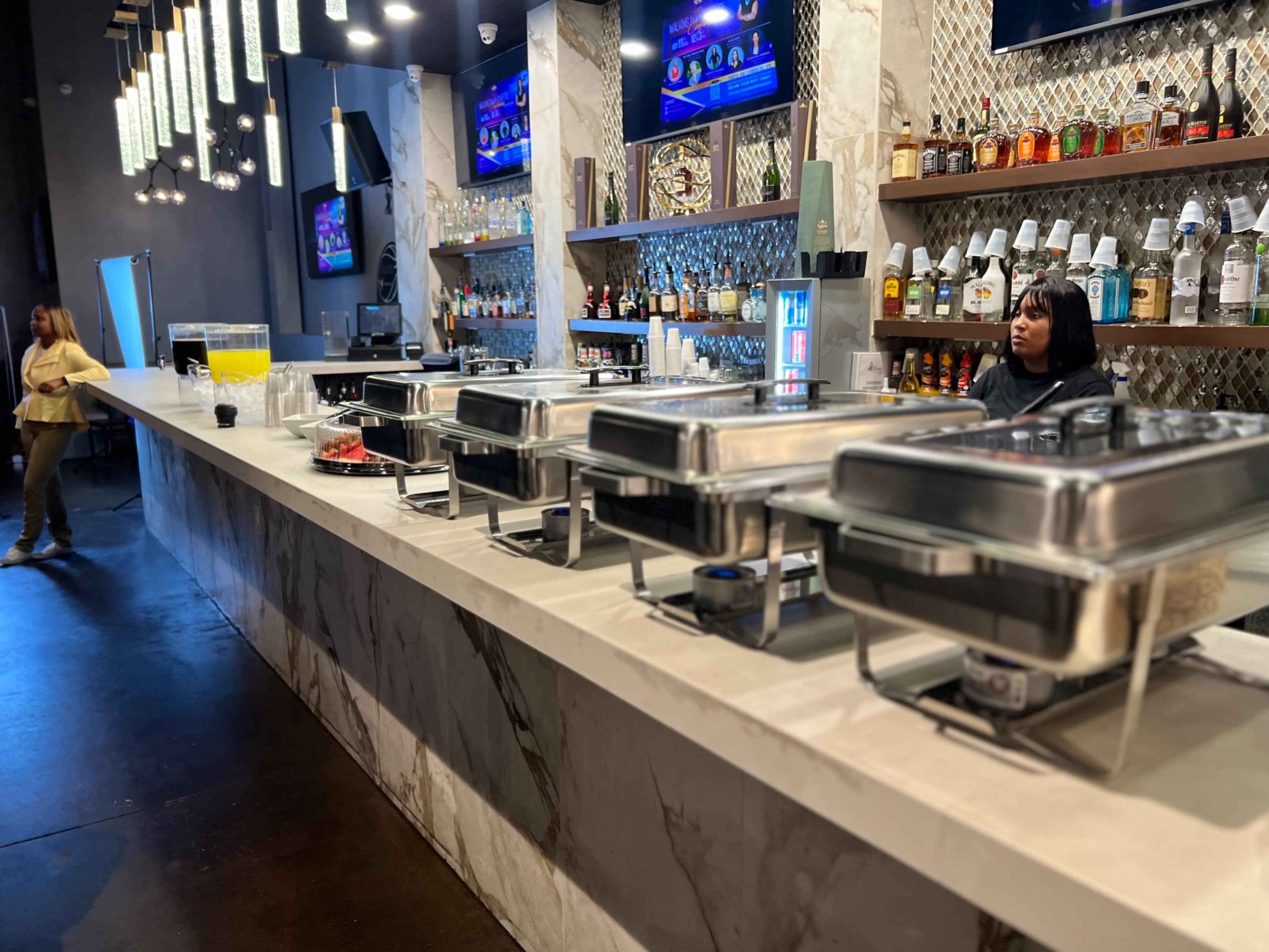 A bar counter with several food trays and a bartender standing behind it, while a person walks in the background.