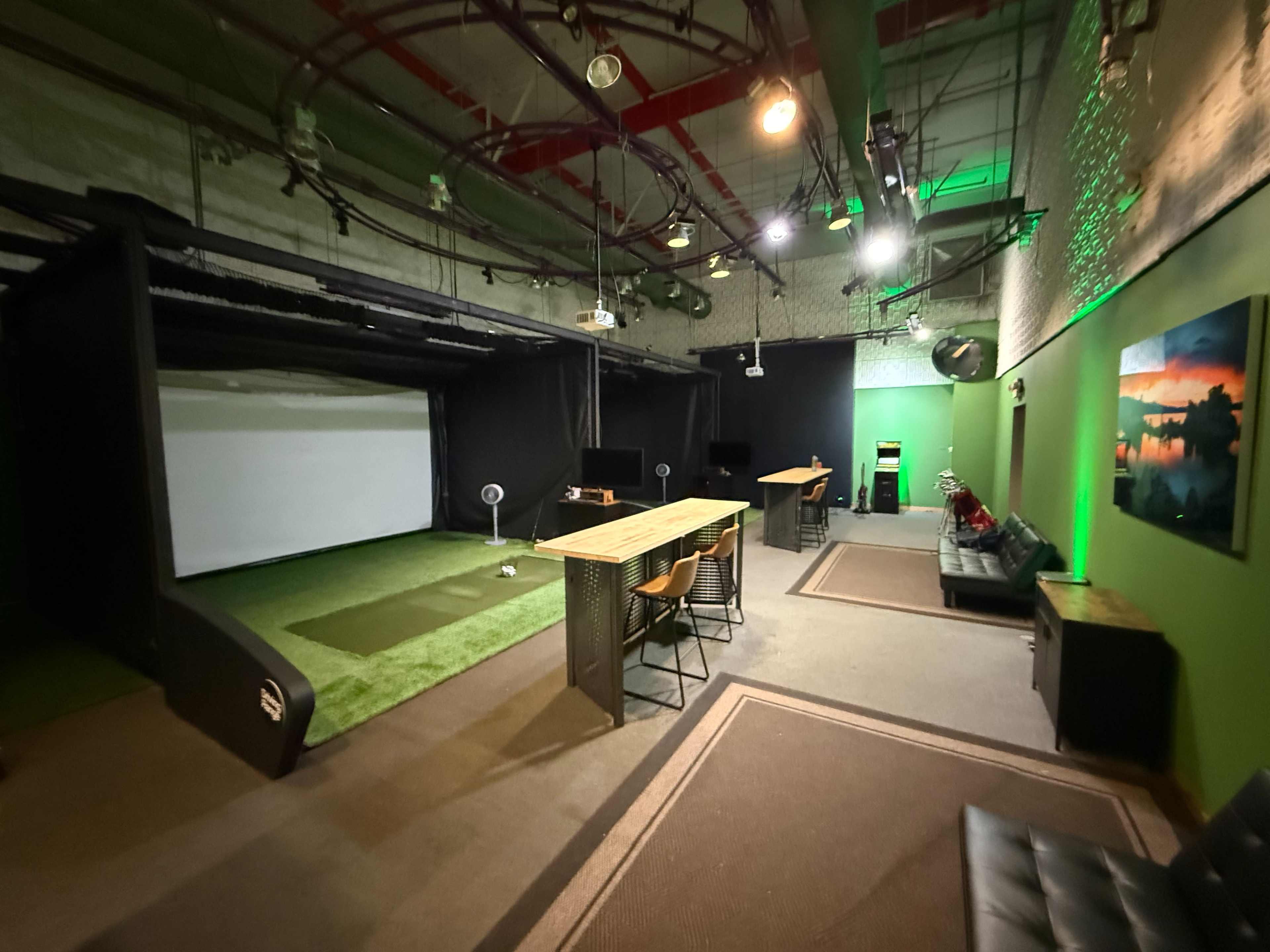 The image depicts an indoor golf simulation room featuring a putting green, a large projection screen, seating, and workspace with a long table and chairs.