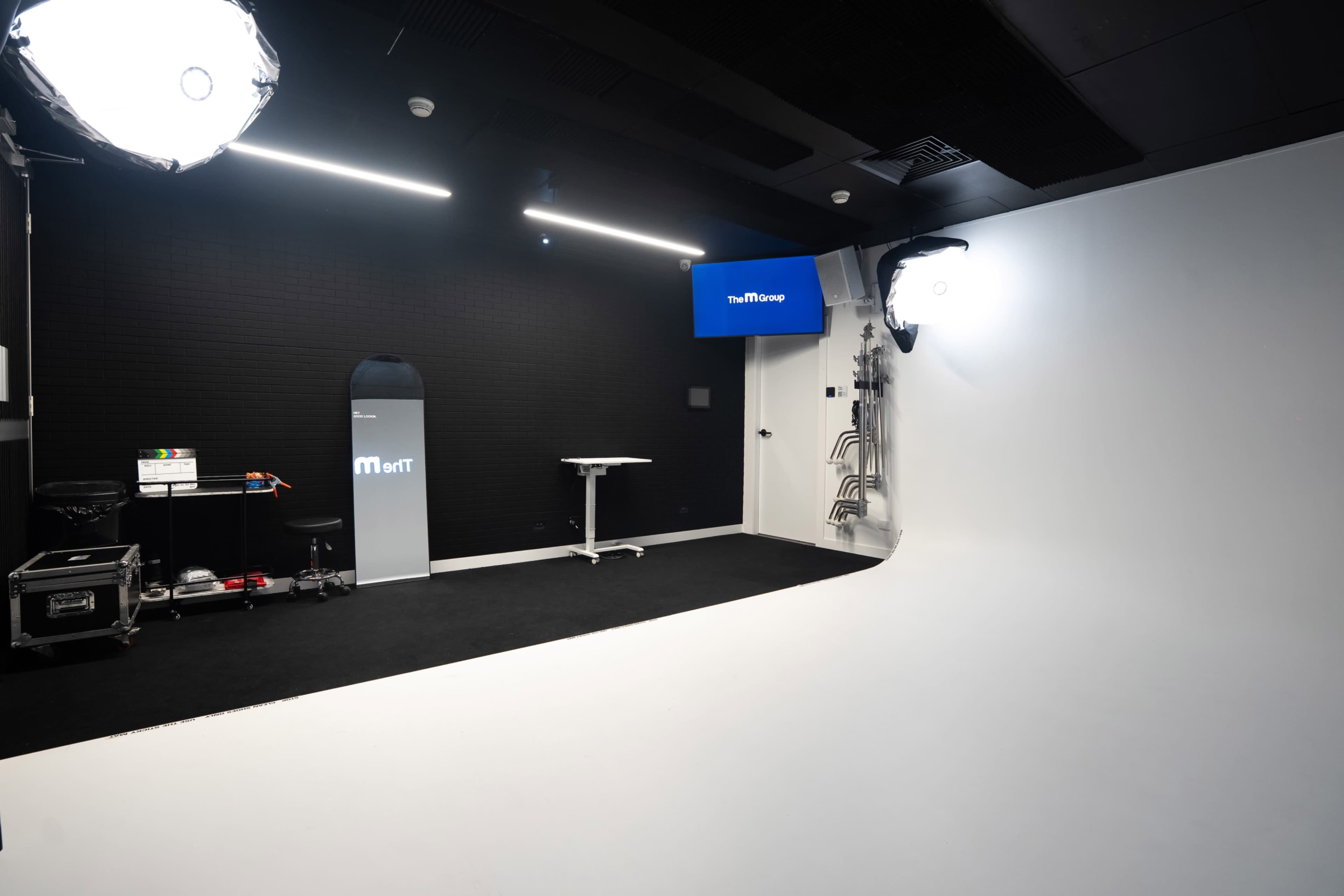 The image shows a clean studio space with a white backdrop, a black floor, and various lighting equipment arranged along the walls.