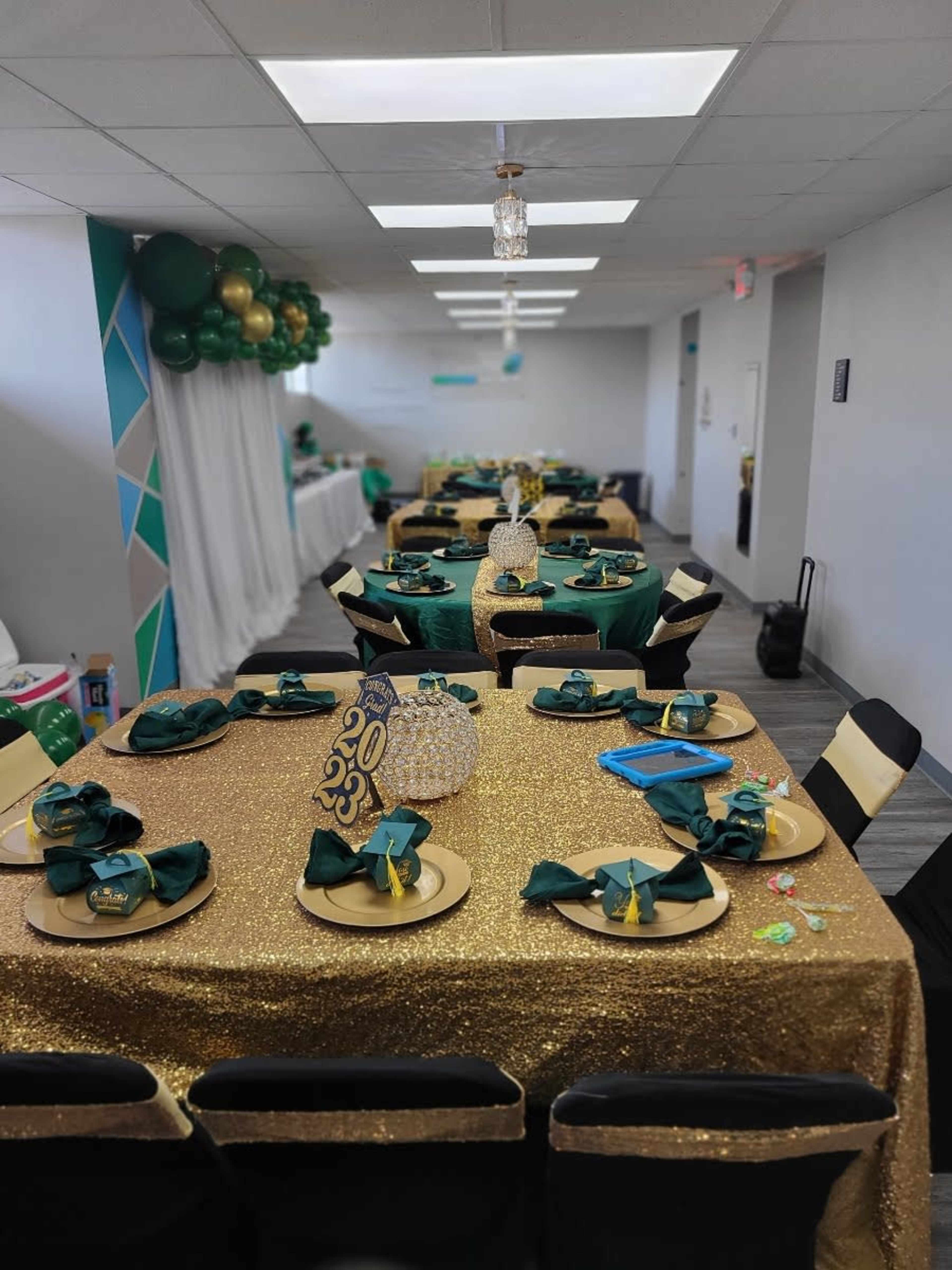 A banquet hall is set with tables covered in gold glitter tablecloths, adorned with green napkins and centerpieces, and decorated with balloons in the background.