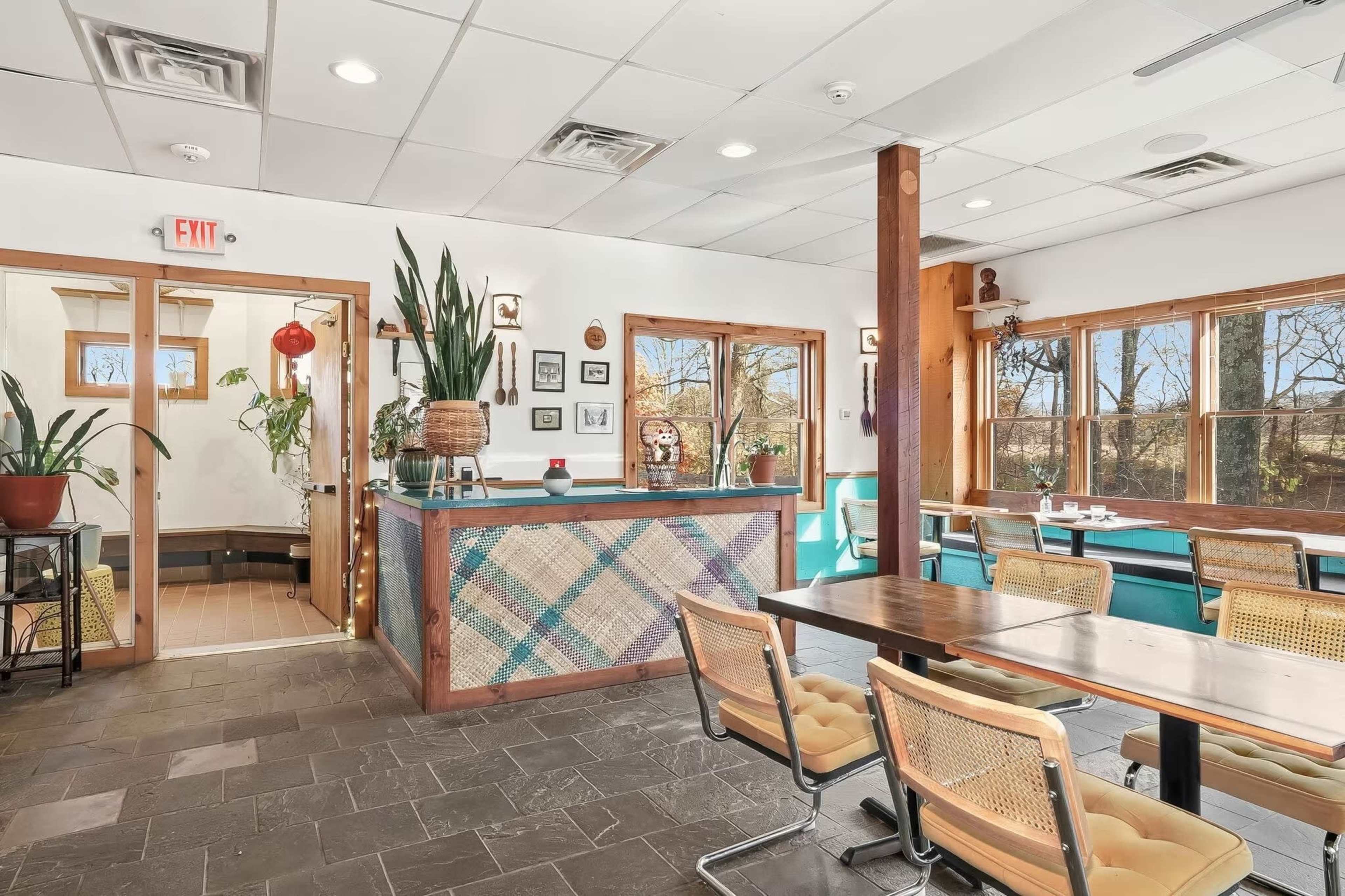 The image shows a brightly lit restaurant interior with wooden accents, large windows, and a reception area featuring plants and decorative items.