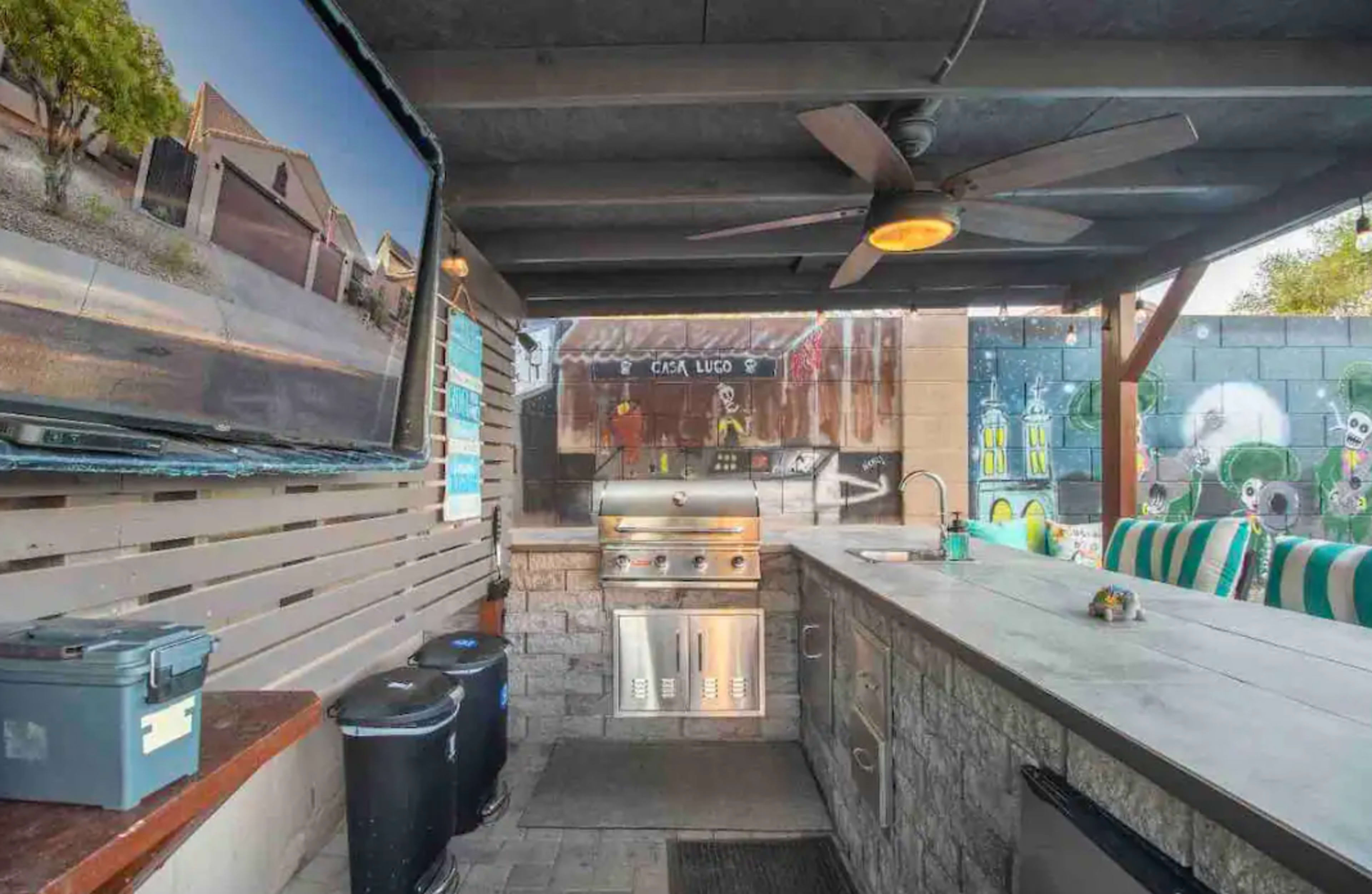 An outdoor kitchen area featuring a grill, counter space, and a mounted TV under a shaded structure.