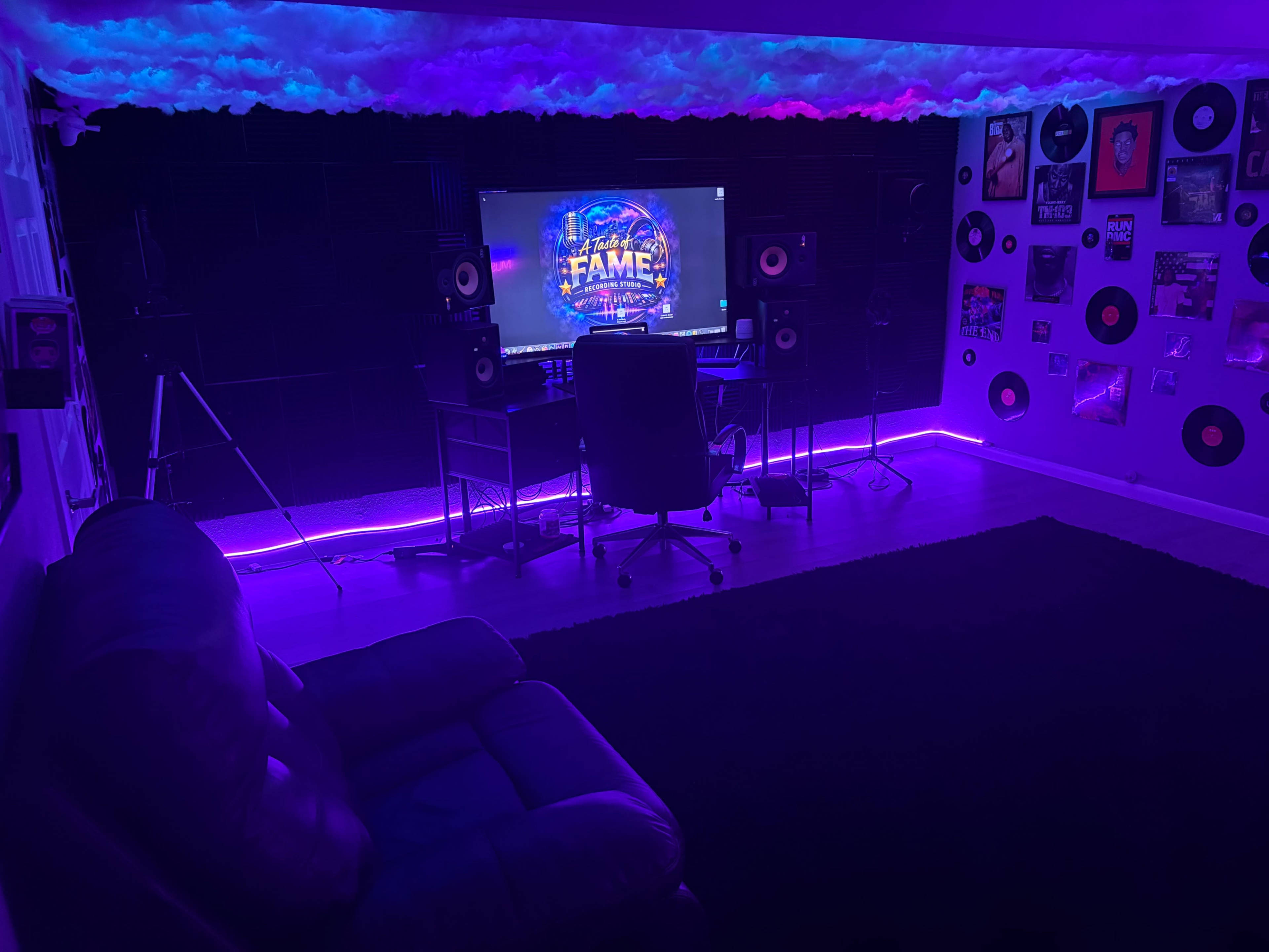 A modern music studio features a desk with a computer, speakers, and a chair, illuminated by purple LED lights with a black rug and a sofa in the corner.