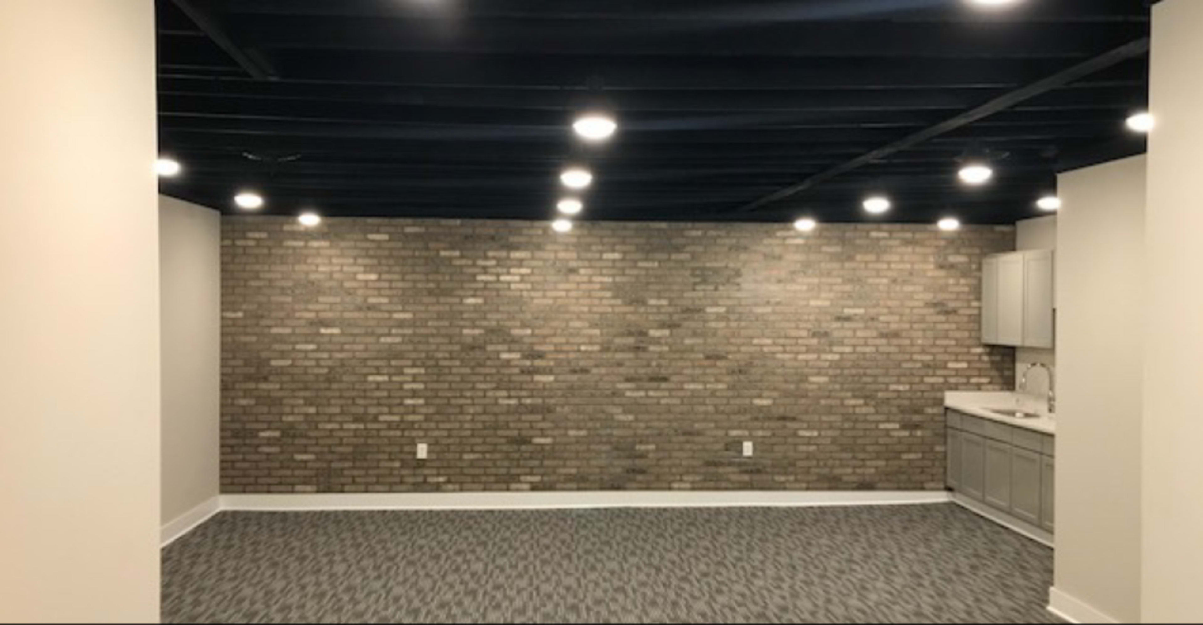 The image shows a room with a brick feature wall, recessed lighting in the ceiling, and a small kitchenette in one corner.