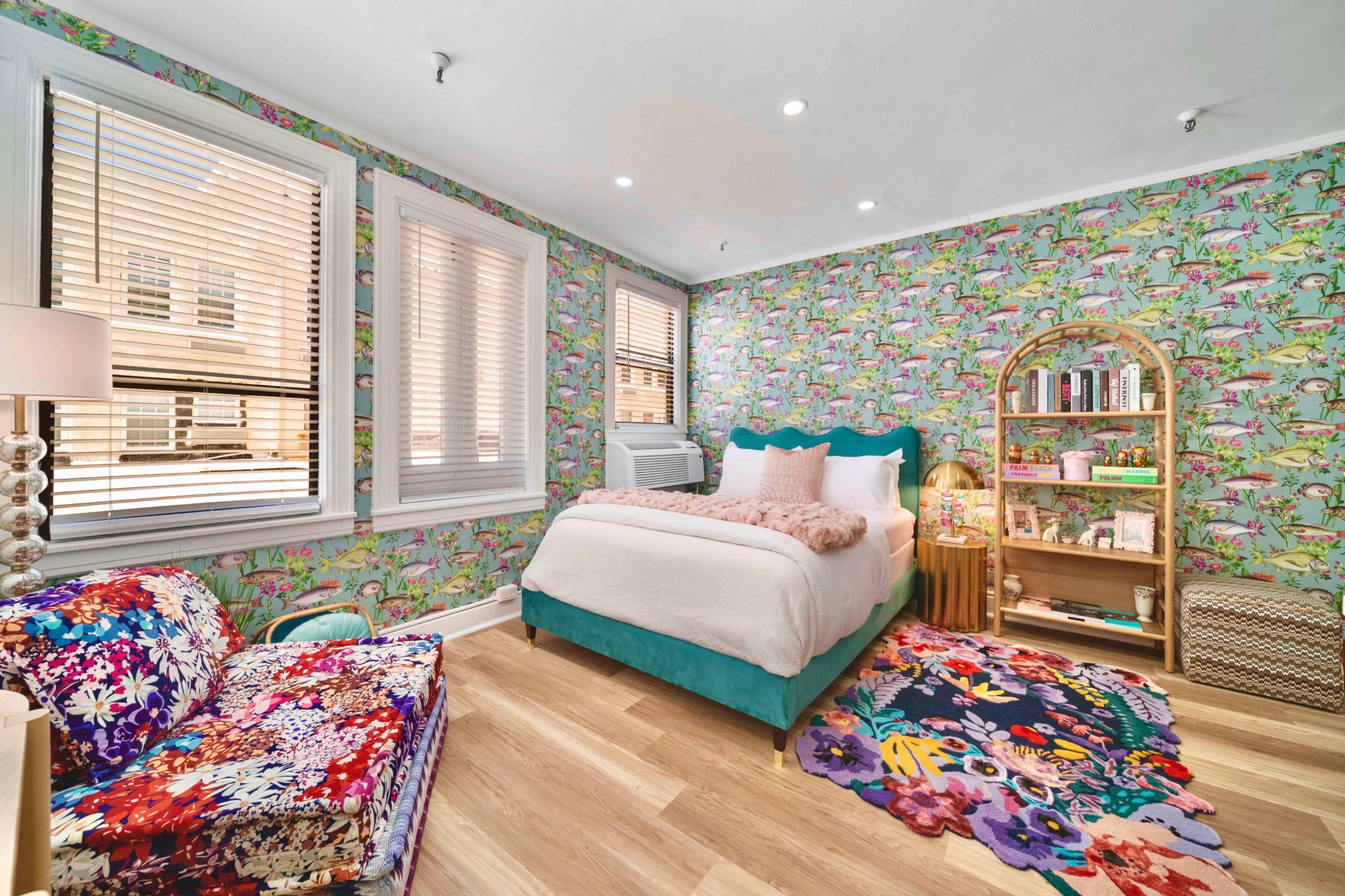 A brightly decorated bedroom with floral wallpaper, a turquoise bed, and a shelf filled with books and decorative items.